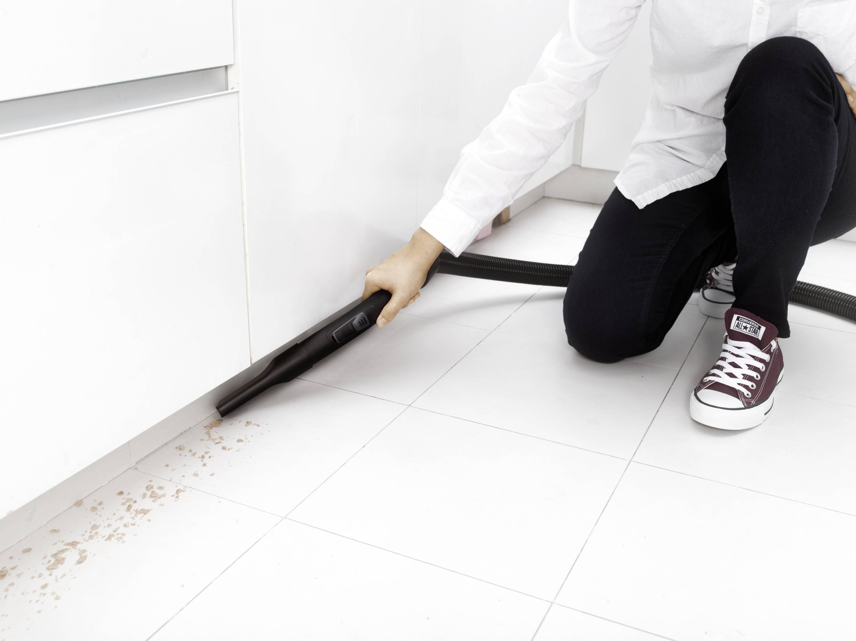 Eine Person in einem weißen Hemd und dunklen Hosen saugt mit einem Staubsauger Krümel vom weißen Küchenboden auf. Sie kniet dabei.