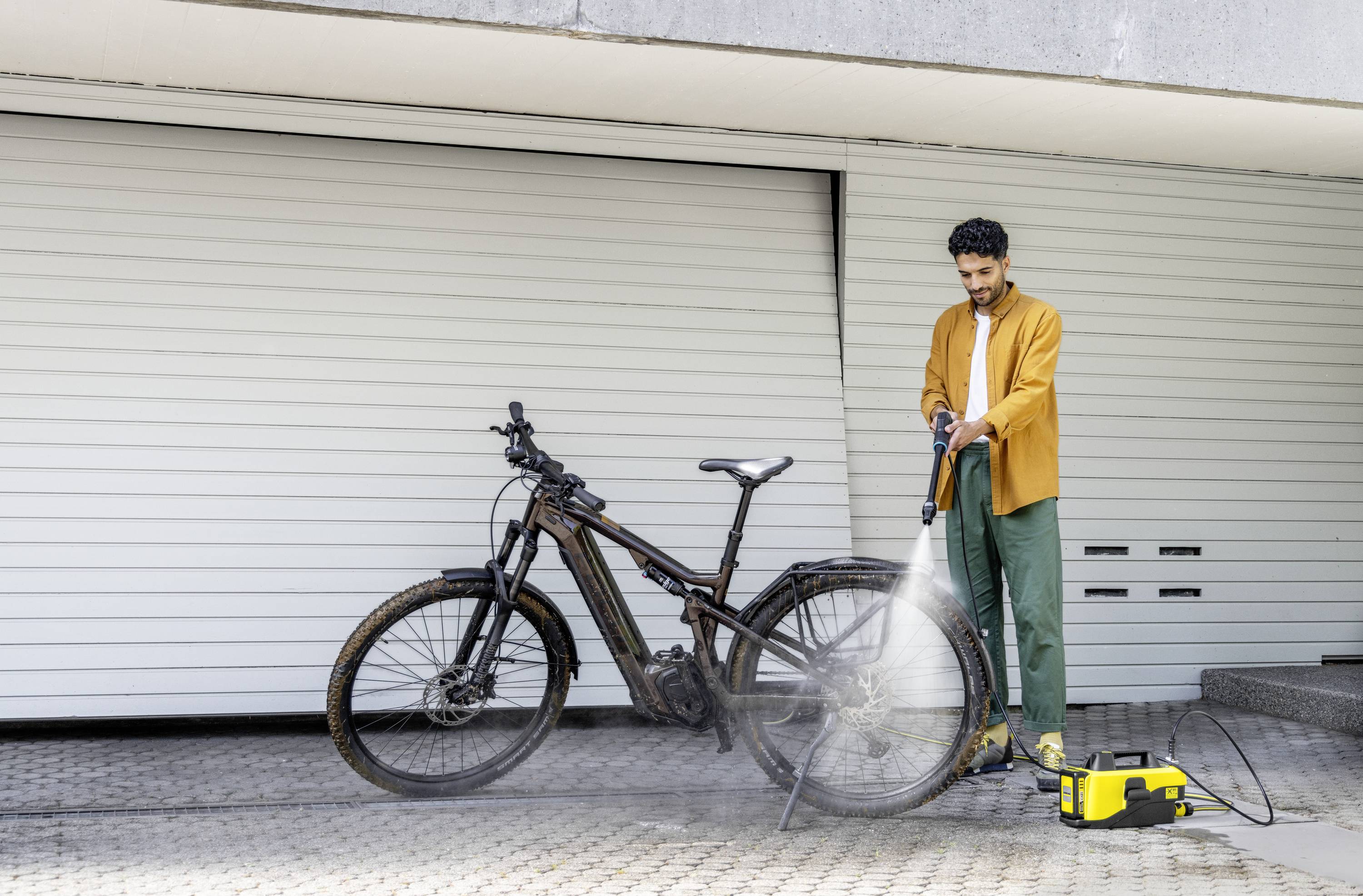 Ein Mann reinigt ein Mountainbike mit einem Hochdruckreiniger vor einem Garagentor.