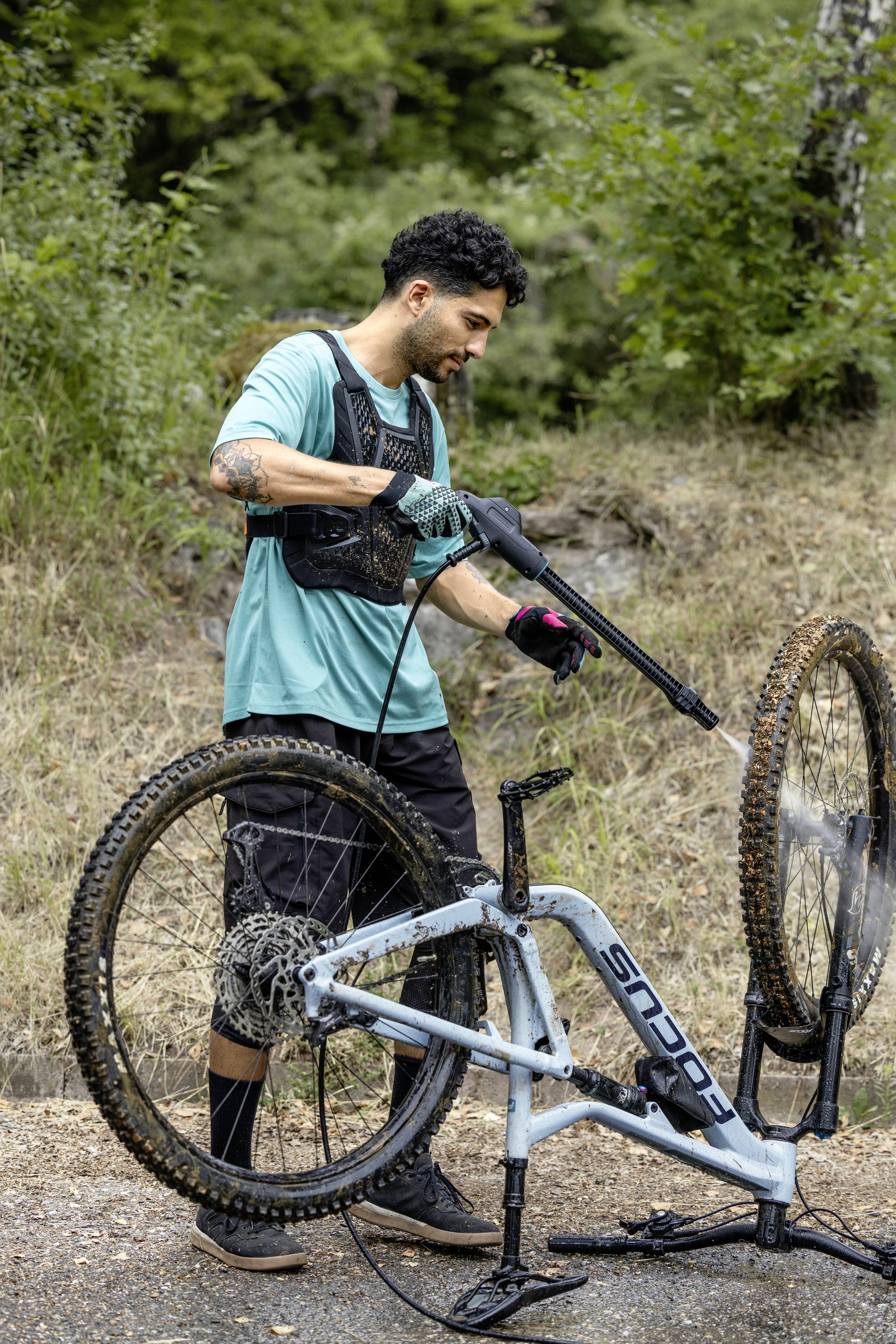 Ein Mann wäscht ein Mountainbike mit einem Hochdruckreiniger auf einem Weg im Wald.
