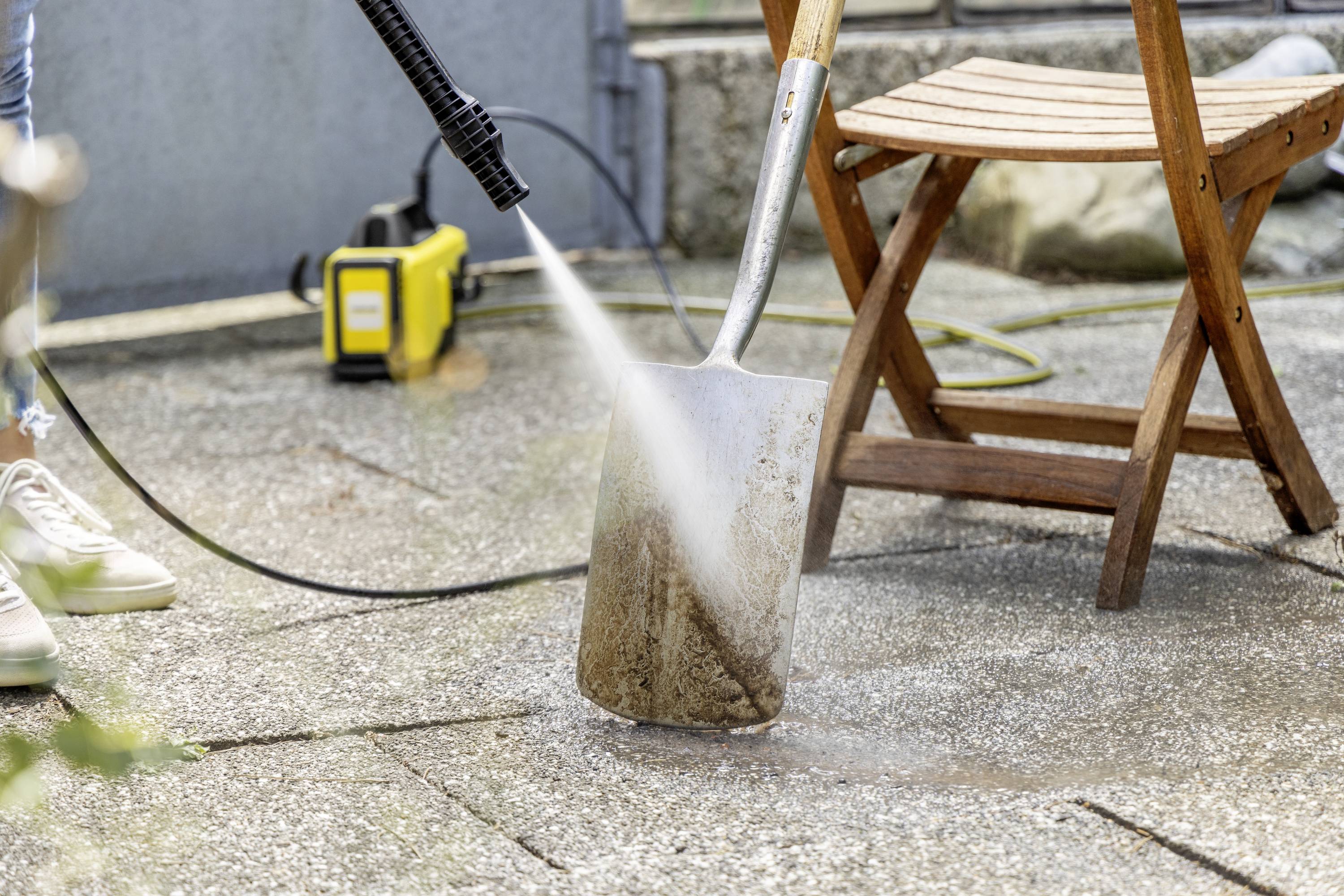 Eine Person reinigt eine Schaufel mit einem Hochdruckreiniger auf einer Terrasse. Im Hintergrund steht ein Holzstuhl.