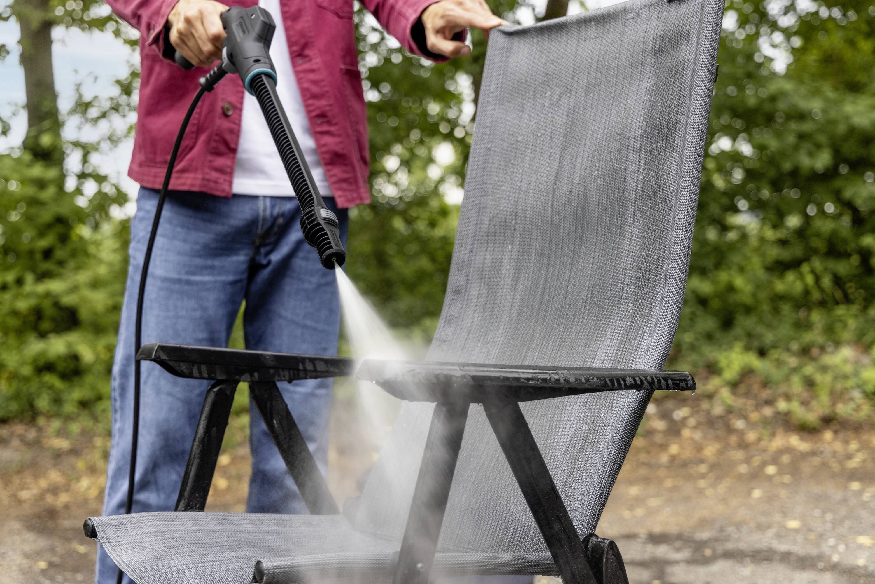 Eine Person reinigt einen grauen Gartenstuhl mit einem Hochdruckreiniger im Freien, im Hintergrund sind Bäume zu sehen.