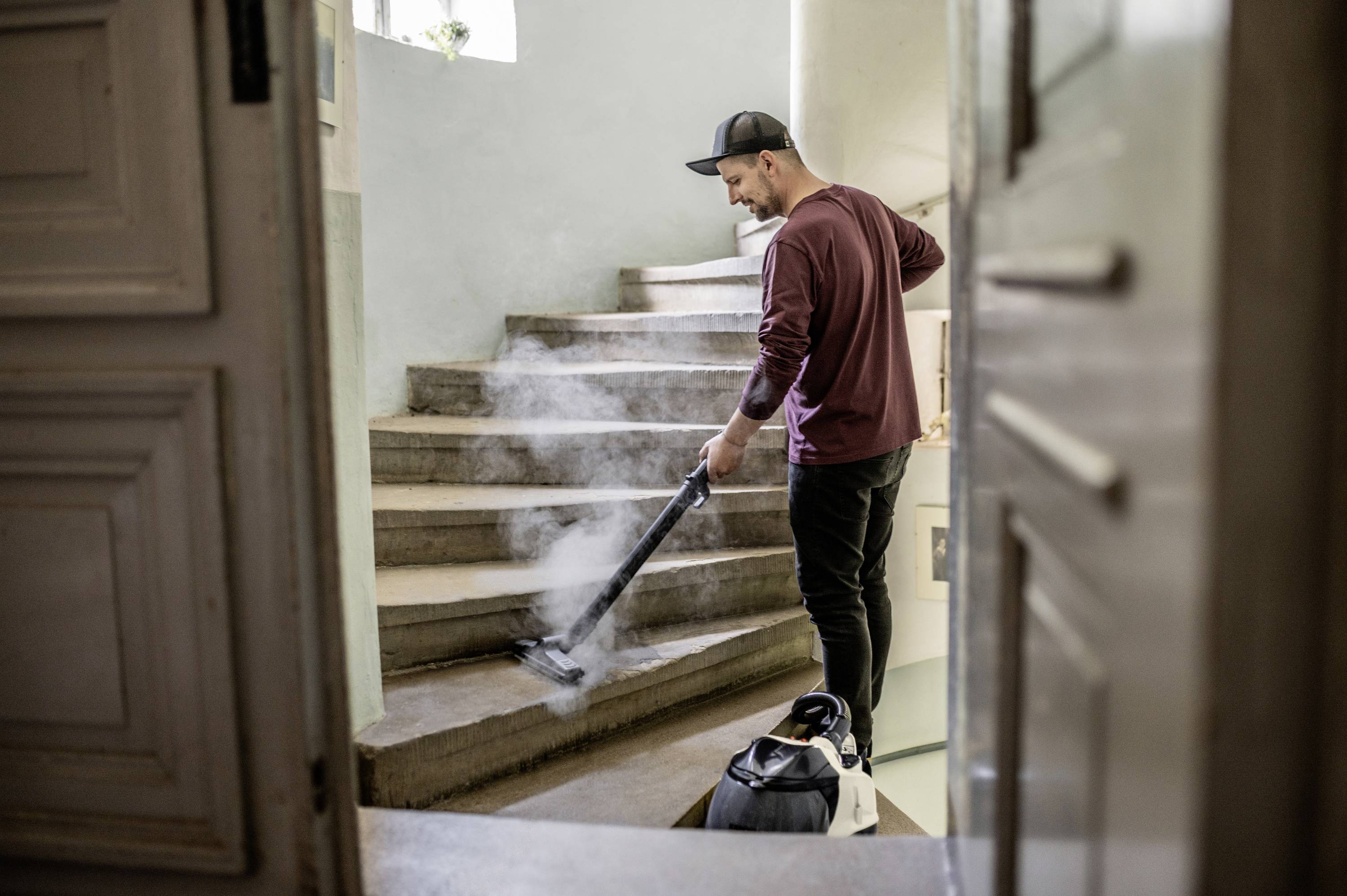 Person reinigt eine Treppe mit einem Dampfsauger. Sie trägt Freizeitkleidung und eine Baseballkappe. Umgebung wirkt häuslich.