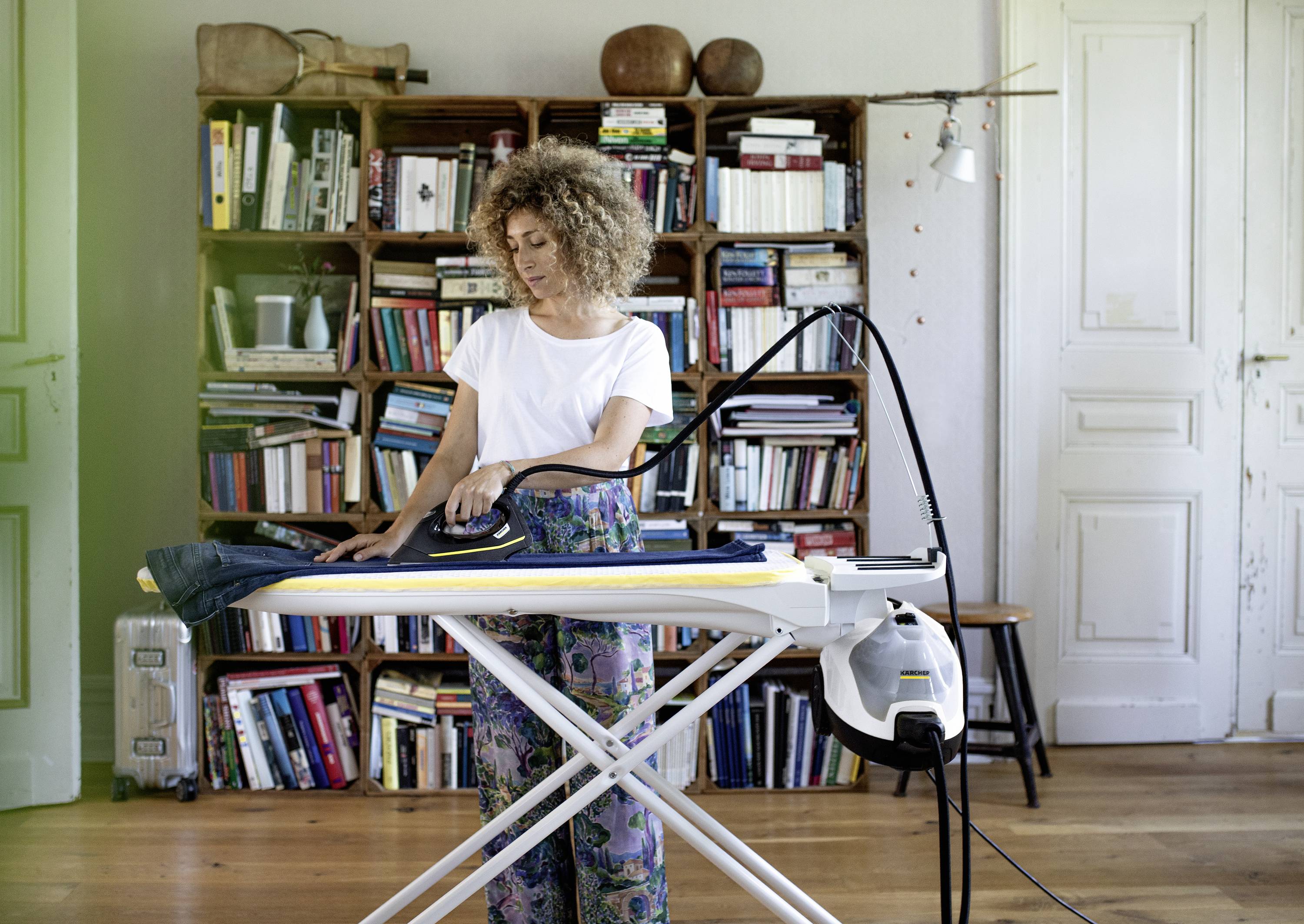 Eine Frau bügelt Kleidung in einem gemütlichen Wohnzimmer mit Büchern im Hintergrund.