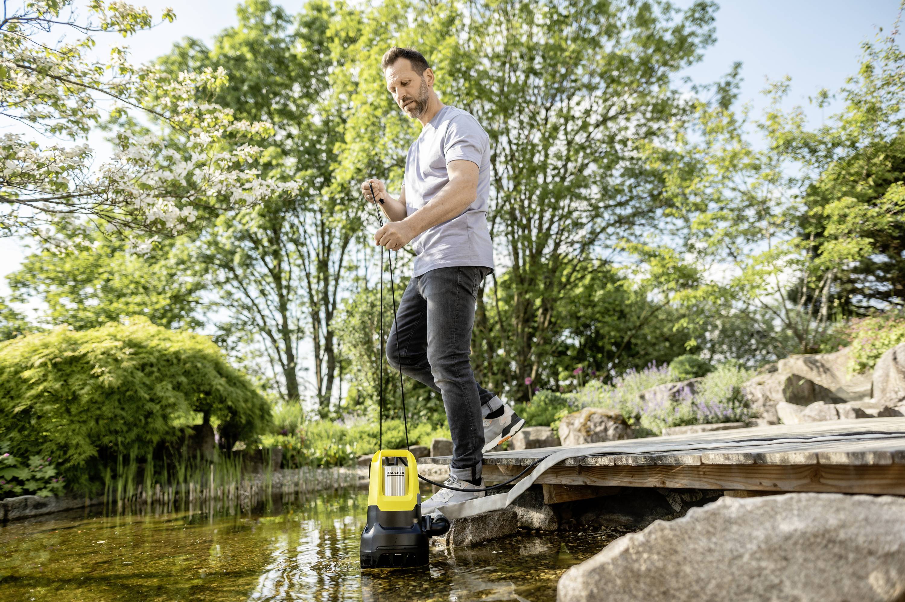 Ein Mann steht auf einem Holzdeck und benutzt eine Tauchpumpe in einem Teich. Der Hintergrund ist ein grüner Garten mit Bäumen.