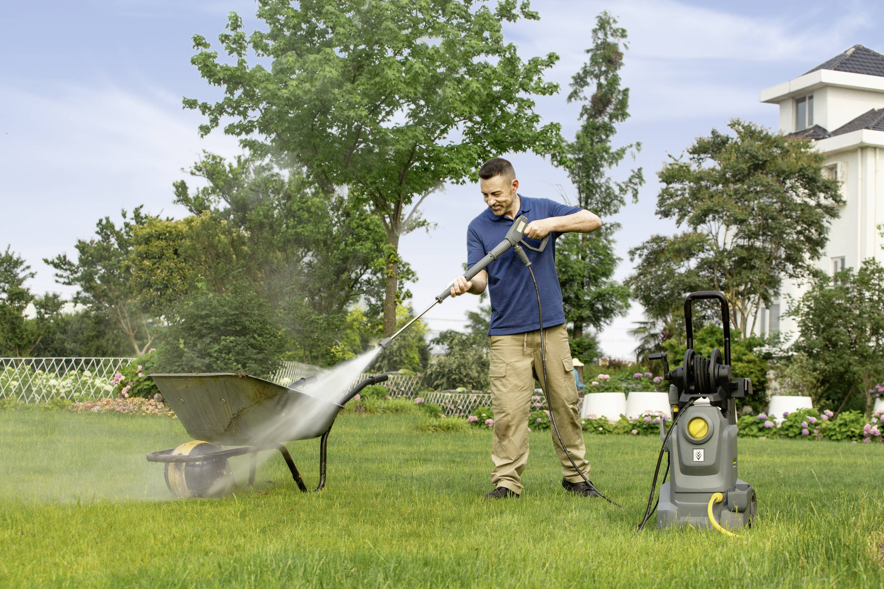 Ein Mann reinigt mit einem Hochdruckreiniger eine Schubkarre im Garten. Im Hintergrund stehen Bäume und ein Wohnhaus.