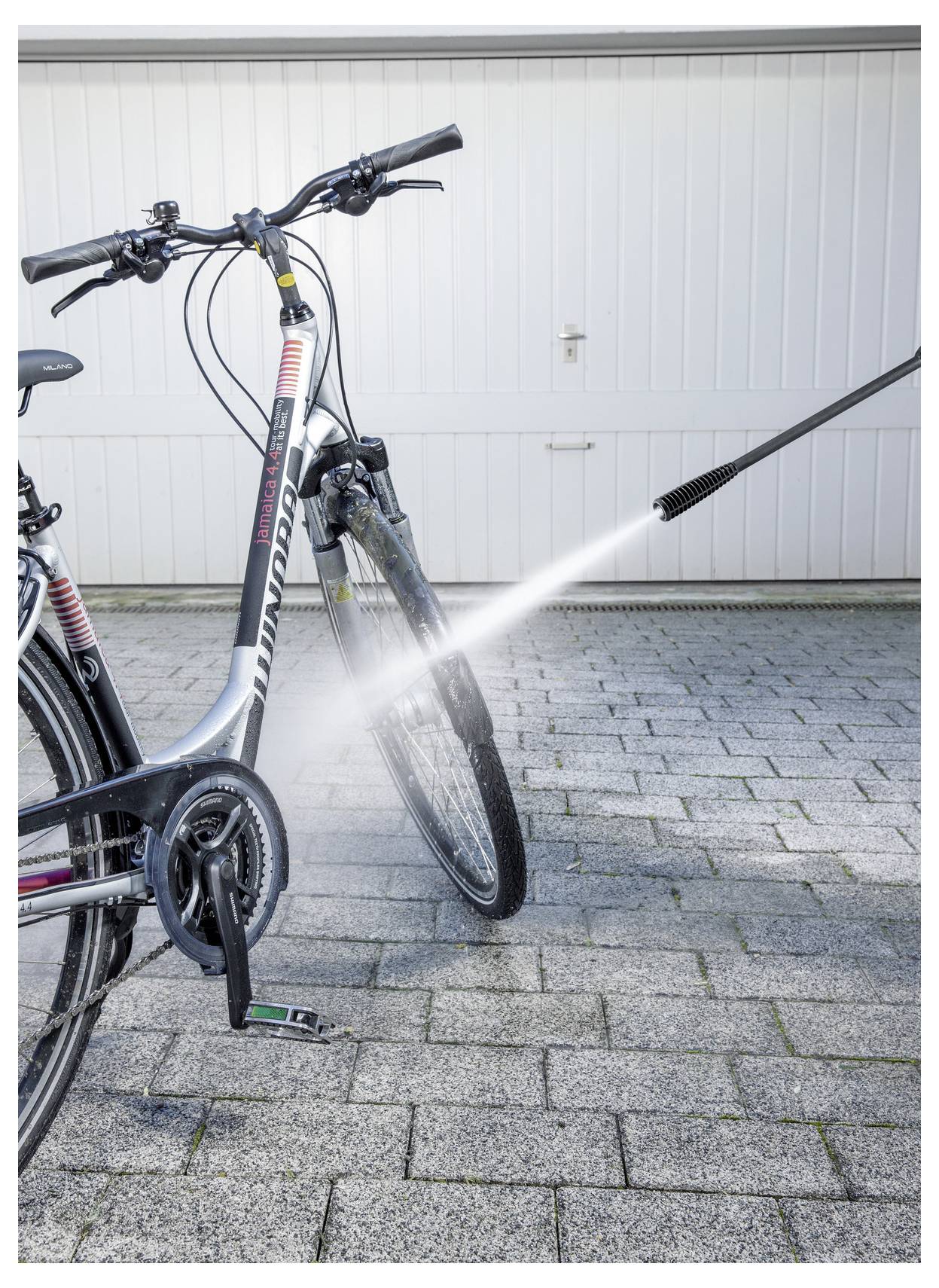 Ein Fahrrad wird mit einem Hochdruckreiniger gereinigt, wobei der Fokus auf dem Vorderrad und dem Rahmen liegt, vor dem Hintergrund eines Garagentors.