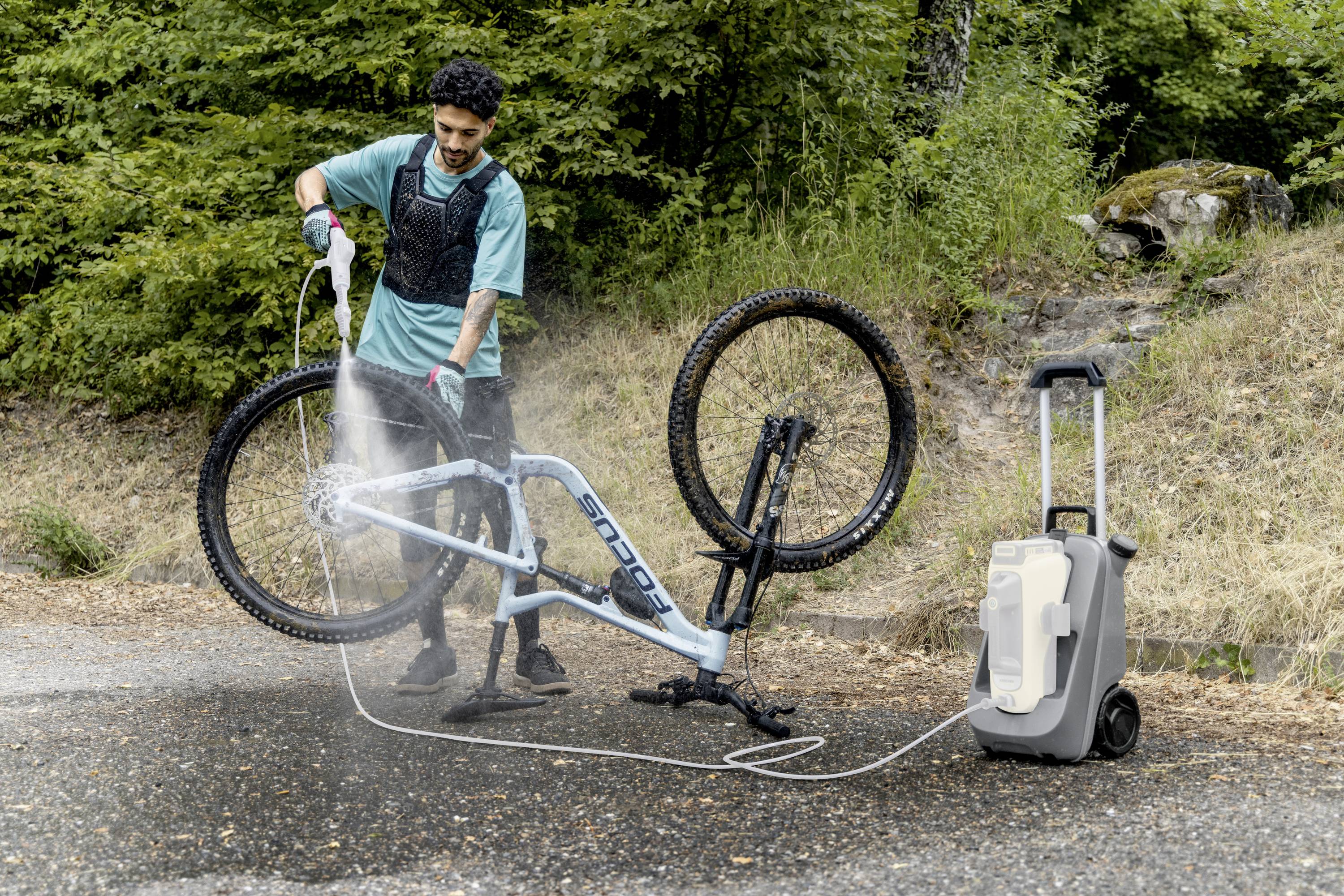 Eine Person reinigt ein auf dem Kopf stehendes Fahrrad mit einem Hochdruckreiniger im Freien. Im Hintergrund sind Bäume zu sehen.