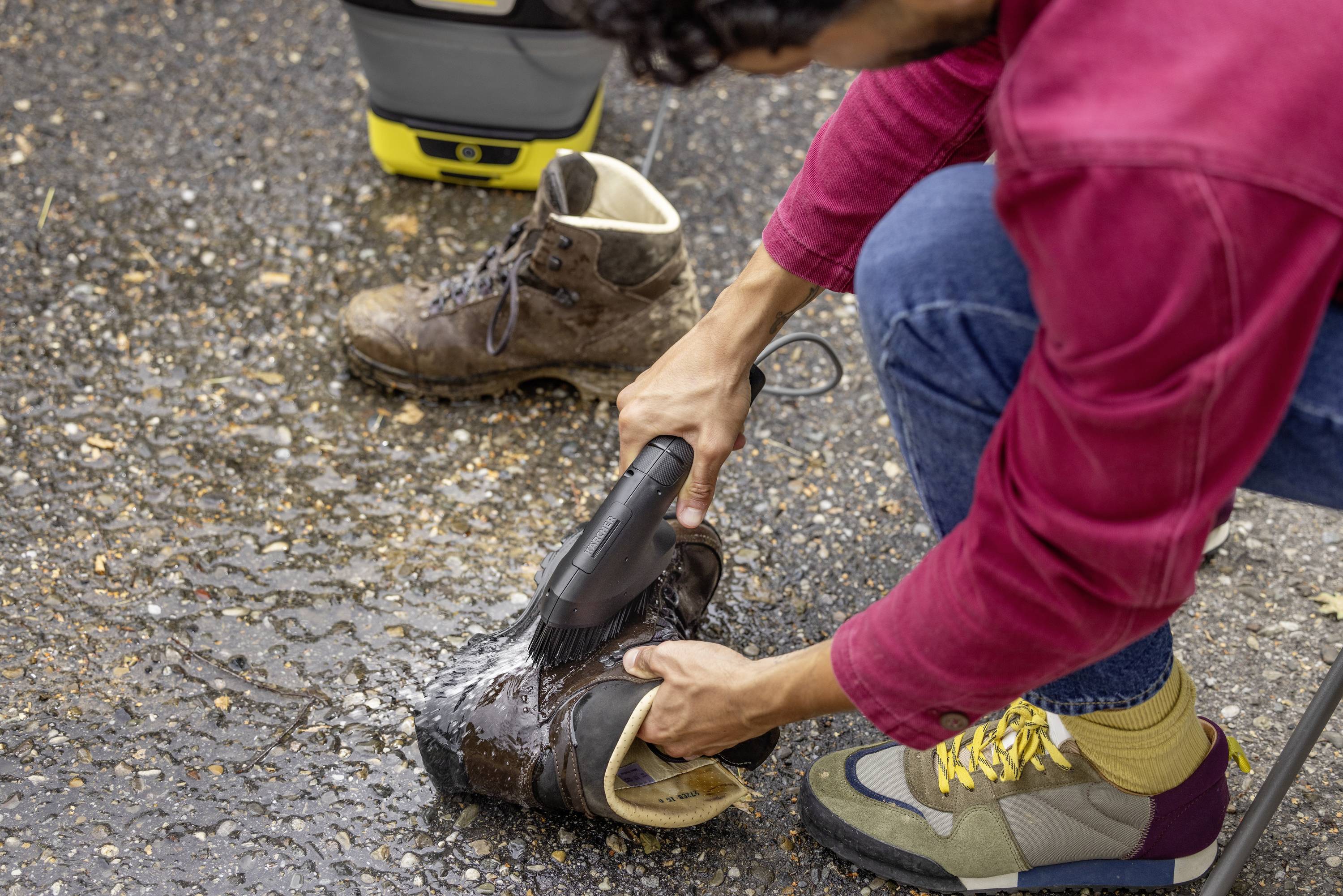 Person reinigt Arbeitsstiefel mit einem Hochdruckreiniger auf Asphalt. Ein weiterer, bereits gereinigter Stiefel steht daneben.