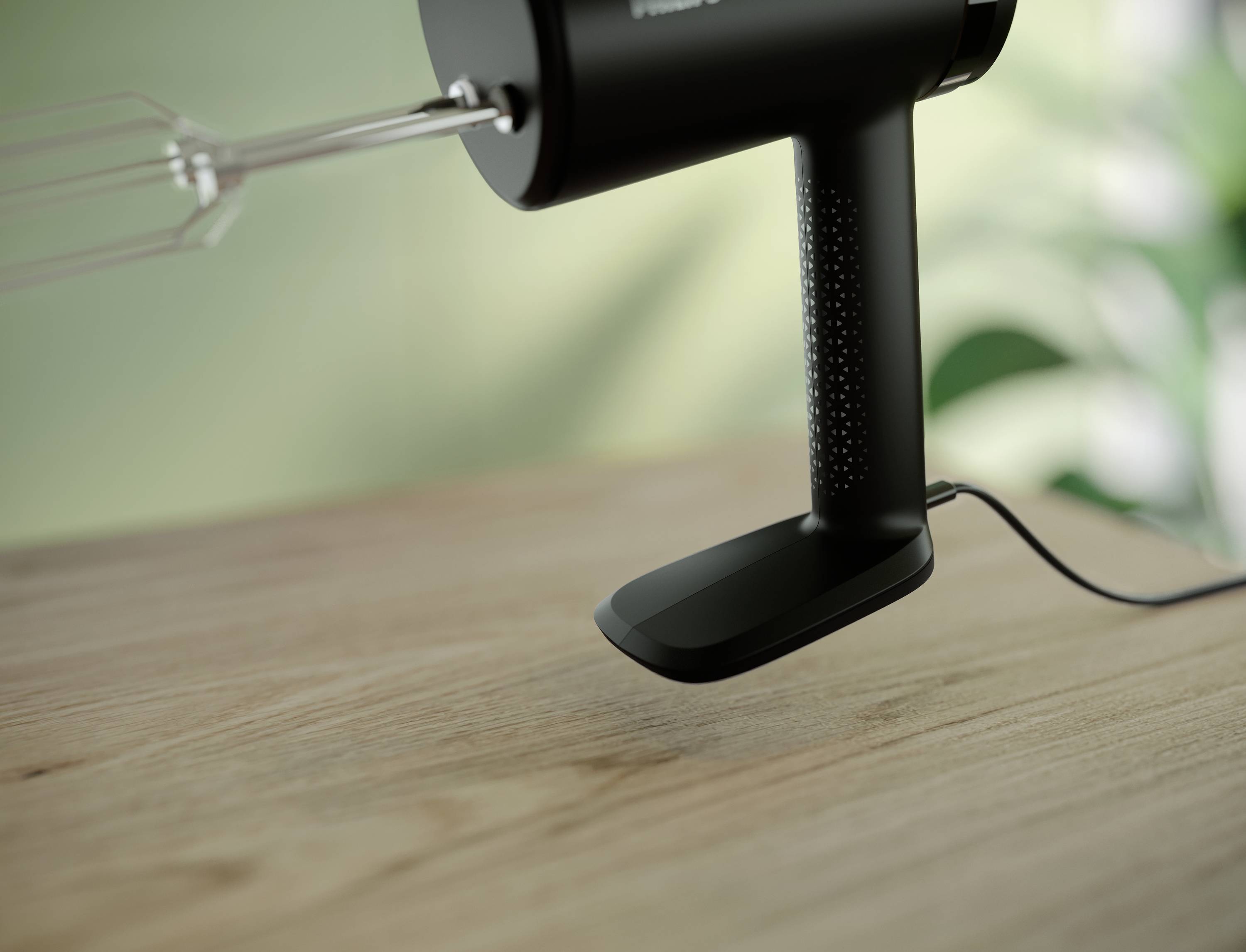 Ein schwarzer Handmixer steht auf einem Holztisch; im Hintergrund sind verschwommene Pflanzen erkennbar.