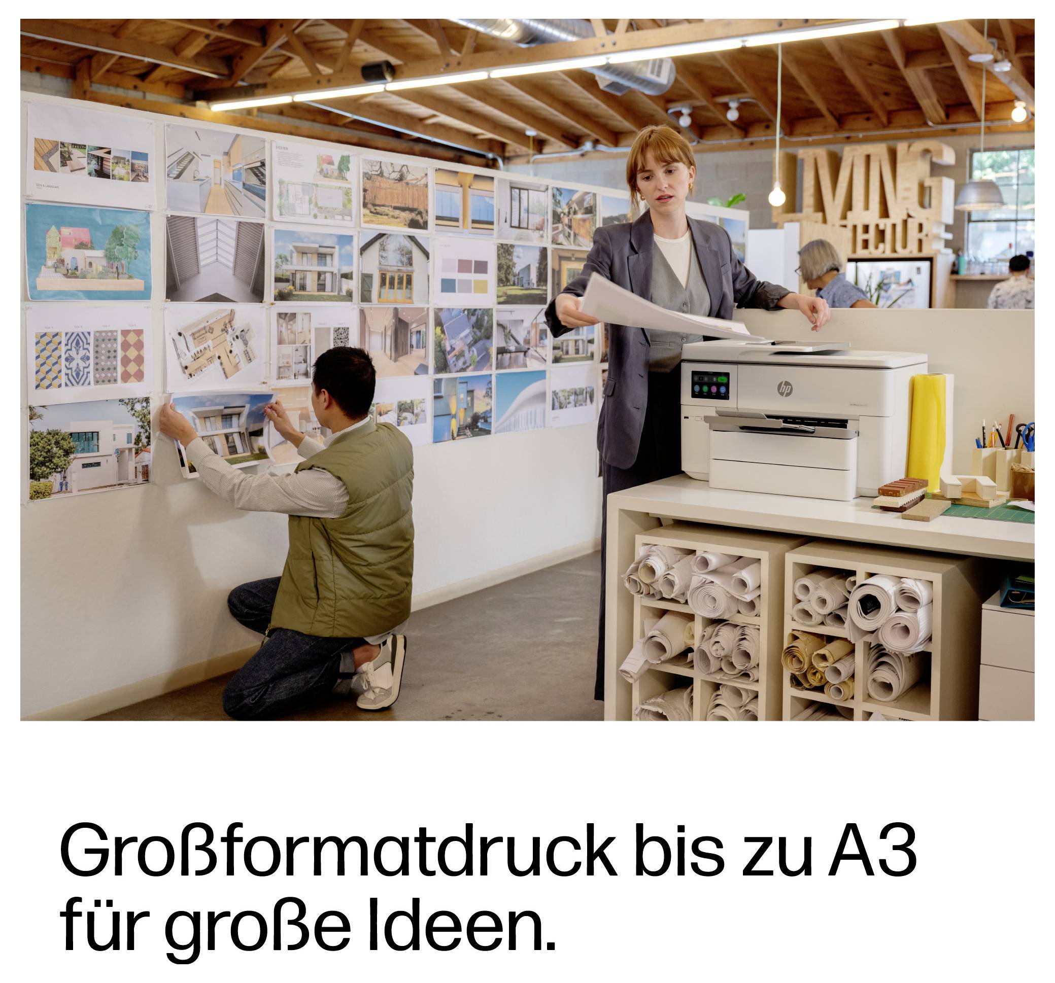 Büroscene mit Menschen, die Designdrucke überprüfen; Drucker im Vordergrund und deutscher Text: 'Großformatdruck bis zu A3 für große Ideen.'