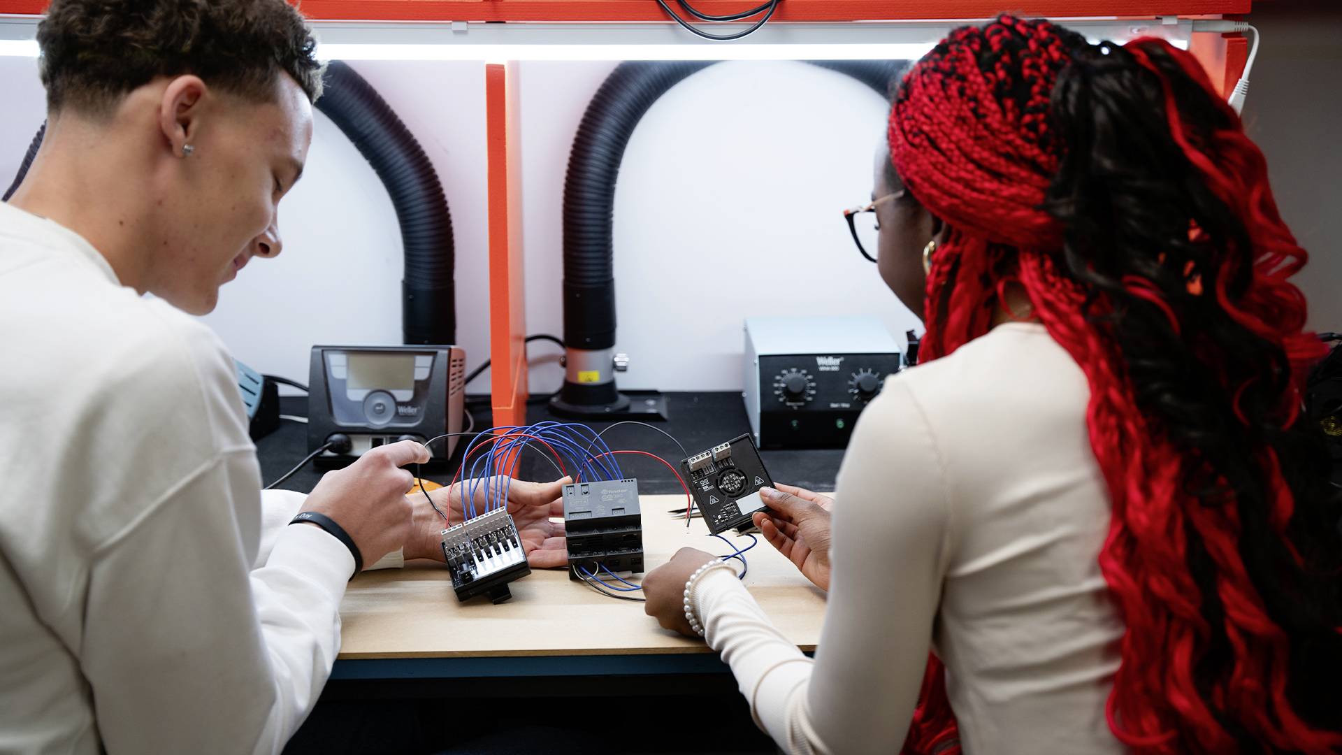 Zwei Personen arbeiten zusammen in einem Elektroniklabor an einem Projekt mit Kabeln und Schaltkreisen auf einem Tisch.