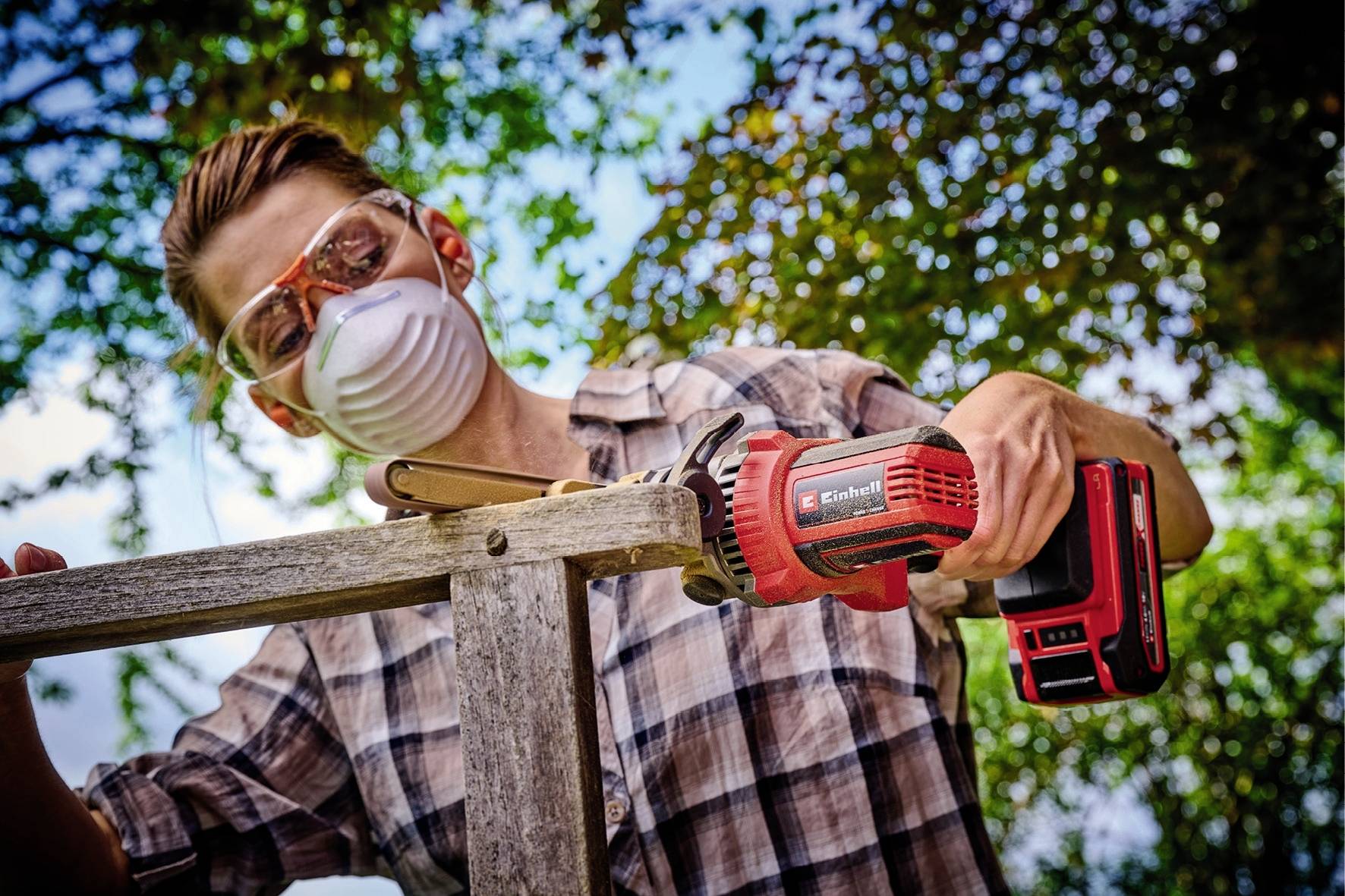 Ein Mann trägt Schutzbrille und Maske, verwendet eine elektrische Säge, um Holz im Freien zu bearbeiten, umgeben von Bäumen.
