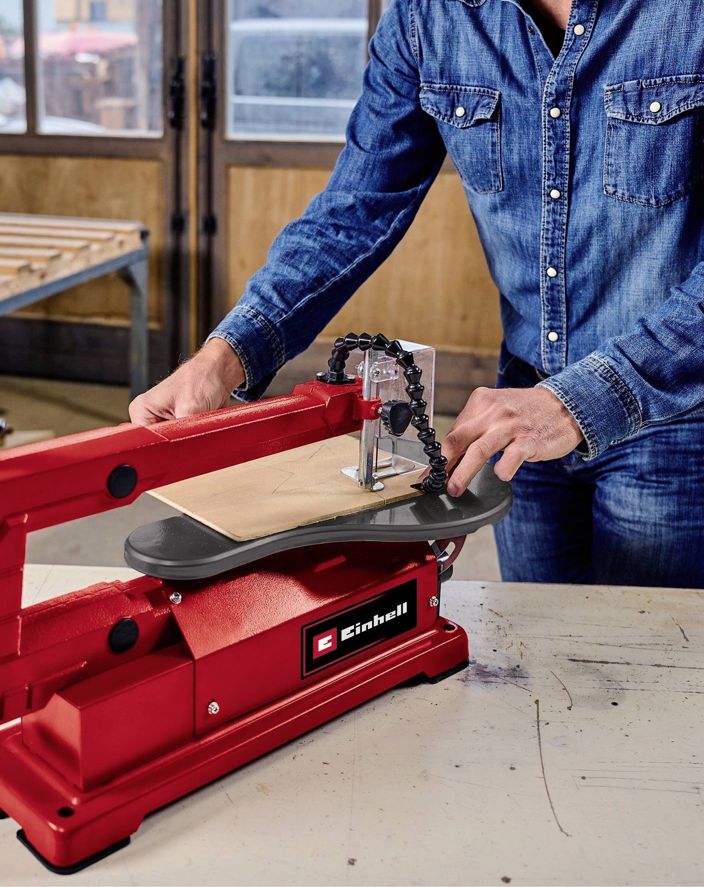 Eine Person in Jeanshemd bedient eine rote Dekupiersäge von Einhell, um ein Holzstück zu schneiden. Die Werkstatt hat Holzwände und Fenster.