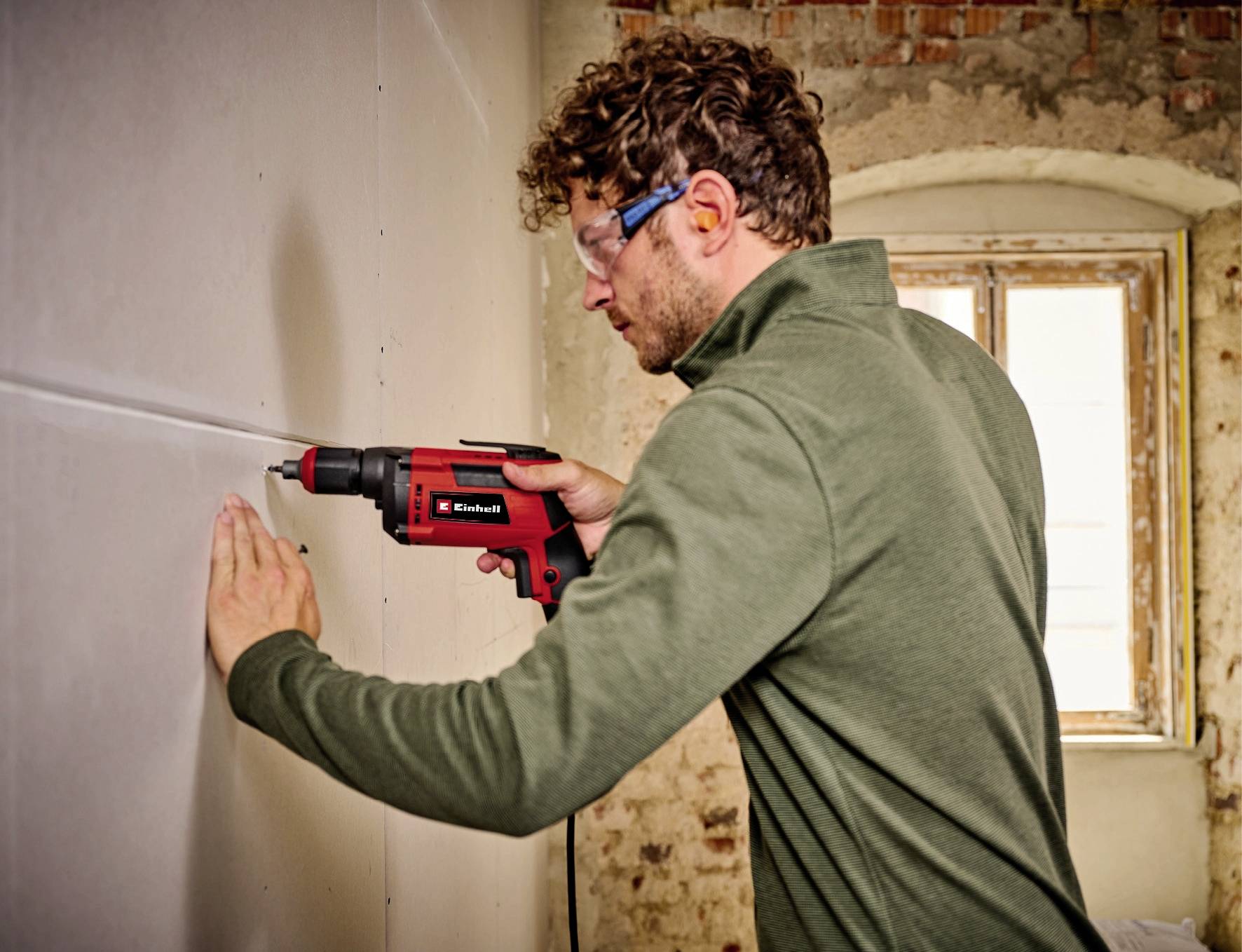 Ein Mann bohrt in eine Wand in einem renovierungsbedürftigen Raum. Er trägt Schutzbrille und benutzt eine rot-schwarze Bohrmaschine.