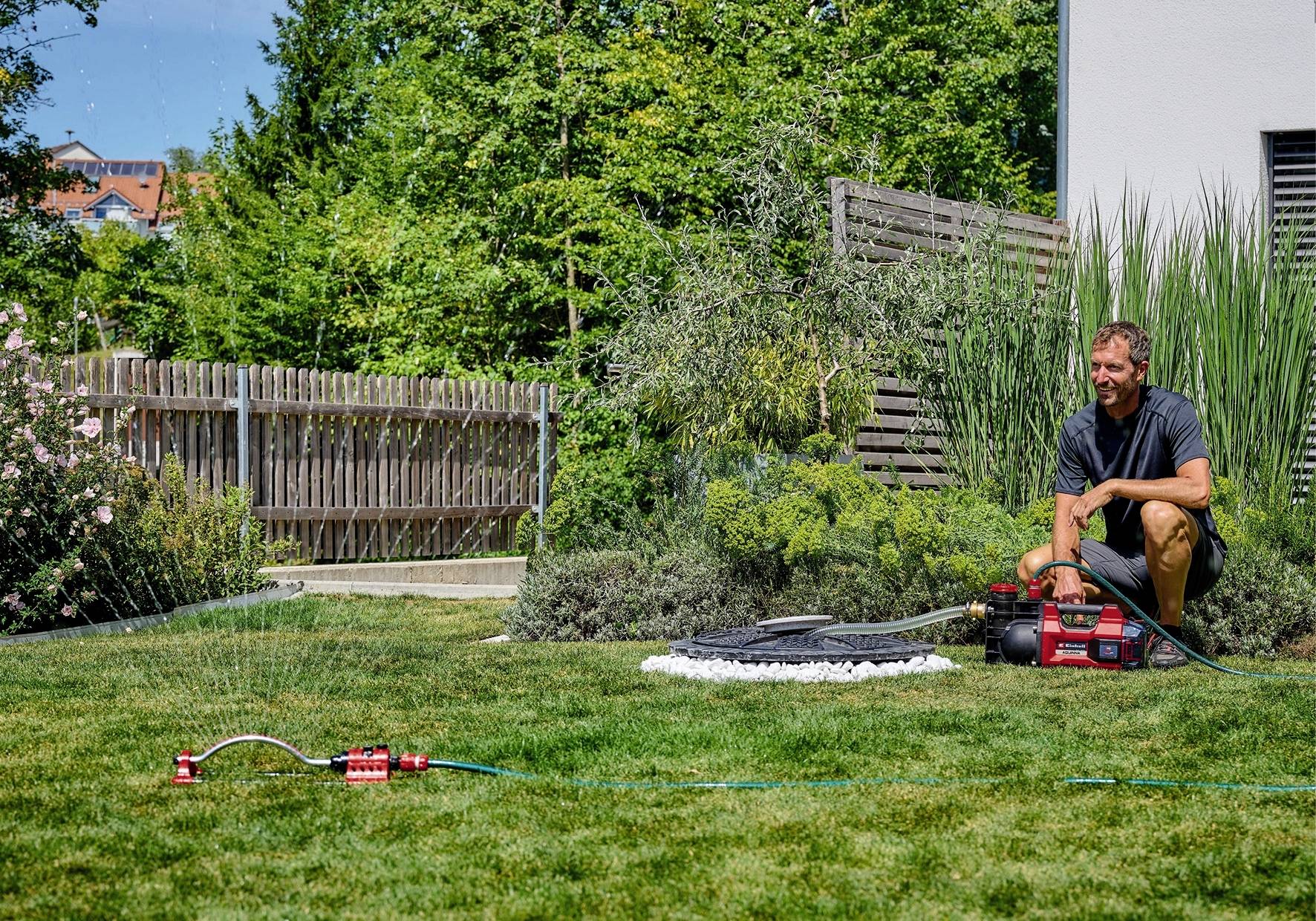 Ein Mann kniet im Garten neben einer Wasserpumpe, während ein Sprinkler das Gras bewässert. Im Hintergrund sind Bäume und ein Zaun.