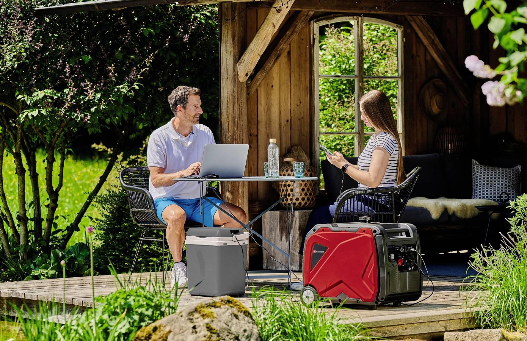 Eine Frau und ein Mann sitzen auf einer Terrasse im Garten und arbeiten an Laptops, ein roter Generator steht nebendran.
