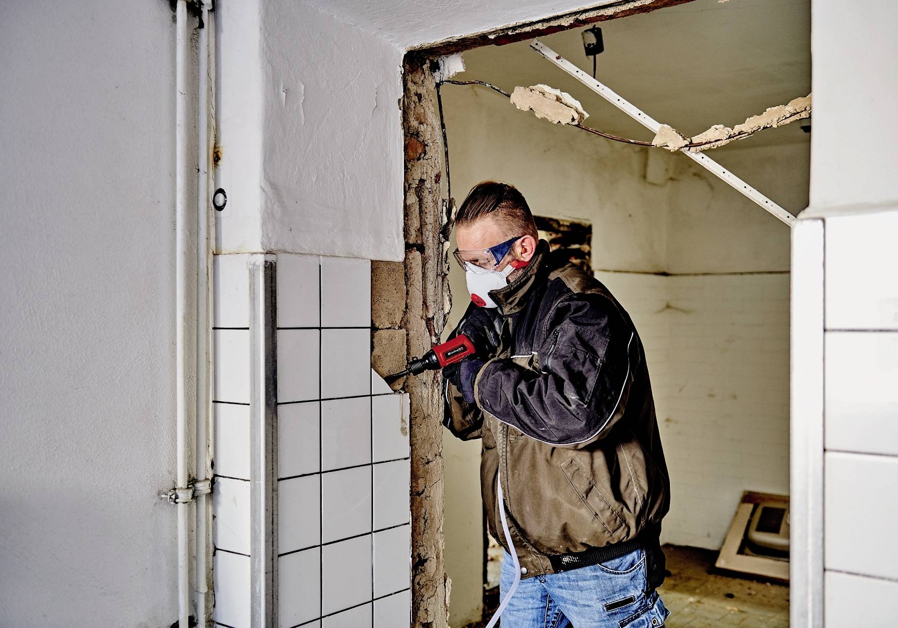 Ein Mann in Schutzkleidung und -brille arbeitet mit einem Bohrhammer an einer Wand in einem teilweise renovierten Raum.