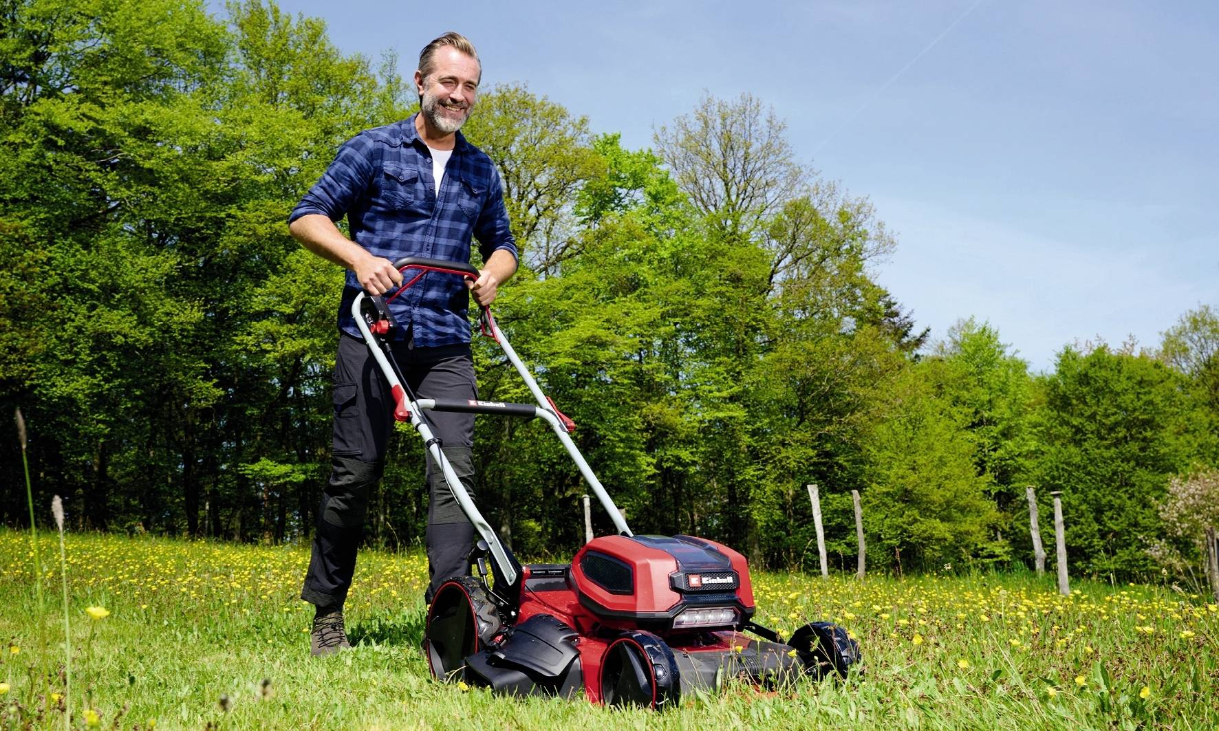 Ein Mann mäht Rasen mit einem elektrischen Rasenmäher auf einer Wiese an einem sonnigen Tag, umgeben von Bäumen.