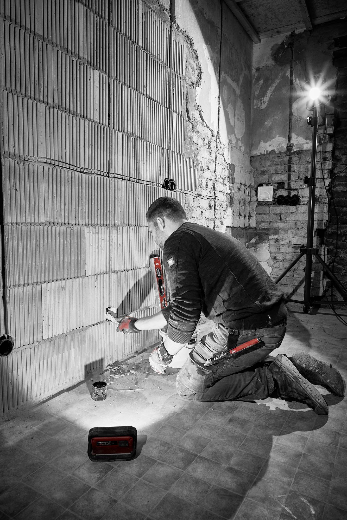 Arbeiter kniet vor einer Wand in einem Rohbau und arbeitet mit Werkzeugen an der Verkabelung. Baustrahler beleuchtet den Raum.