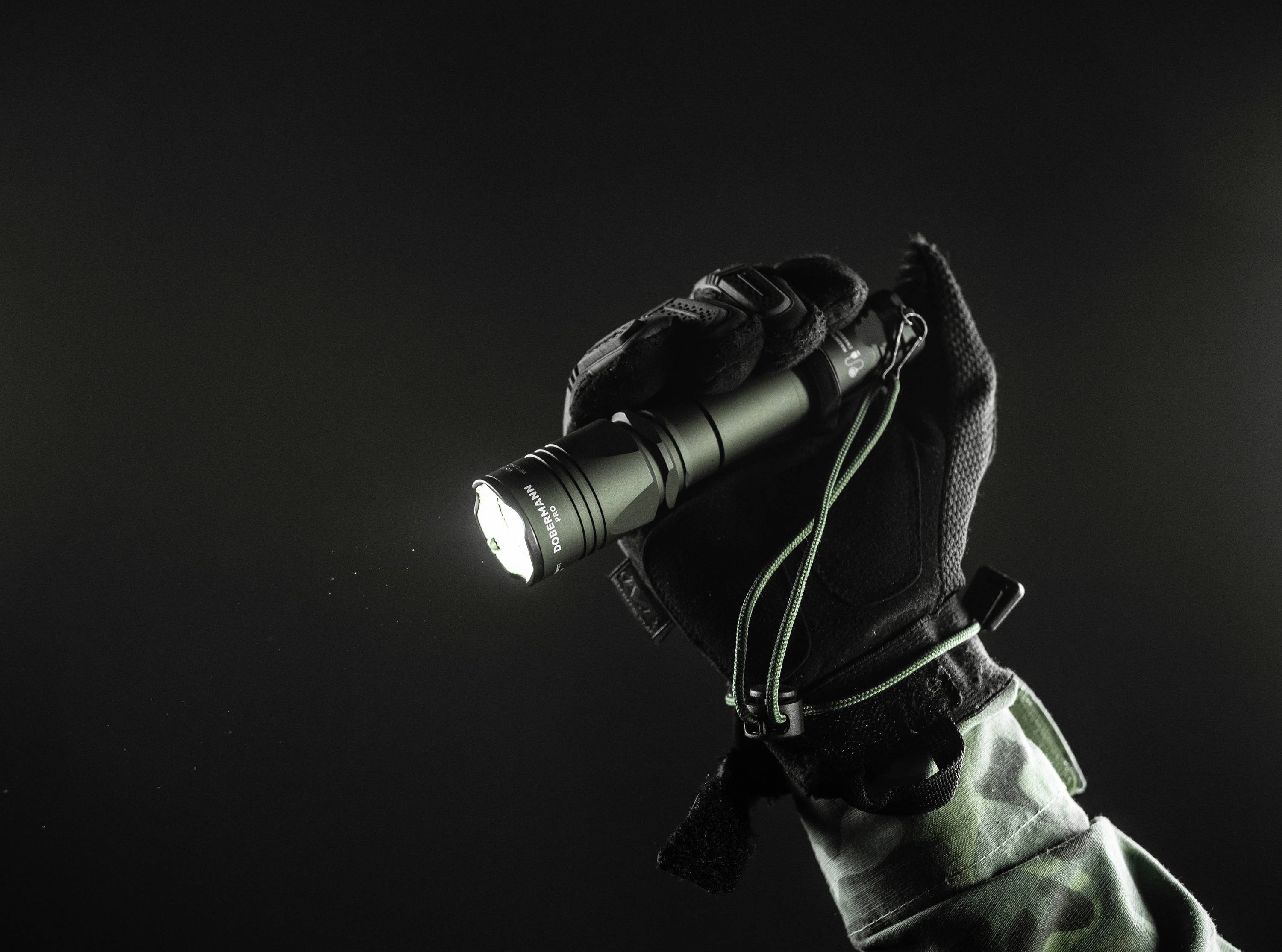 Eine behandschuhte Hand hält eine eingeschaltete Taschenlampe vor einem dunklen Hintergrund, betont durch einen Militärärmel.