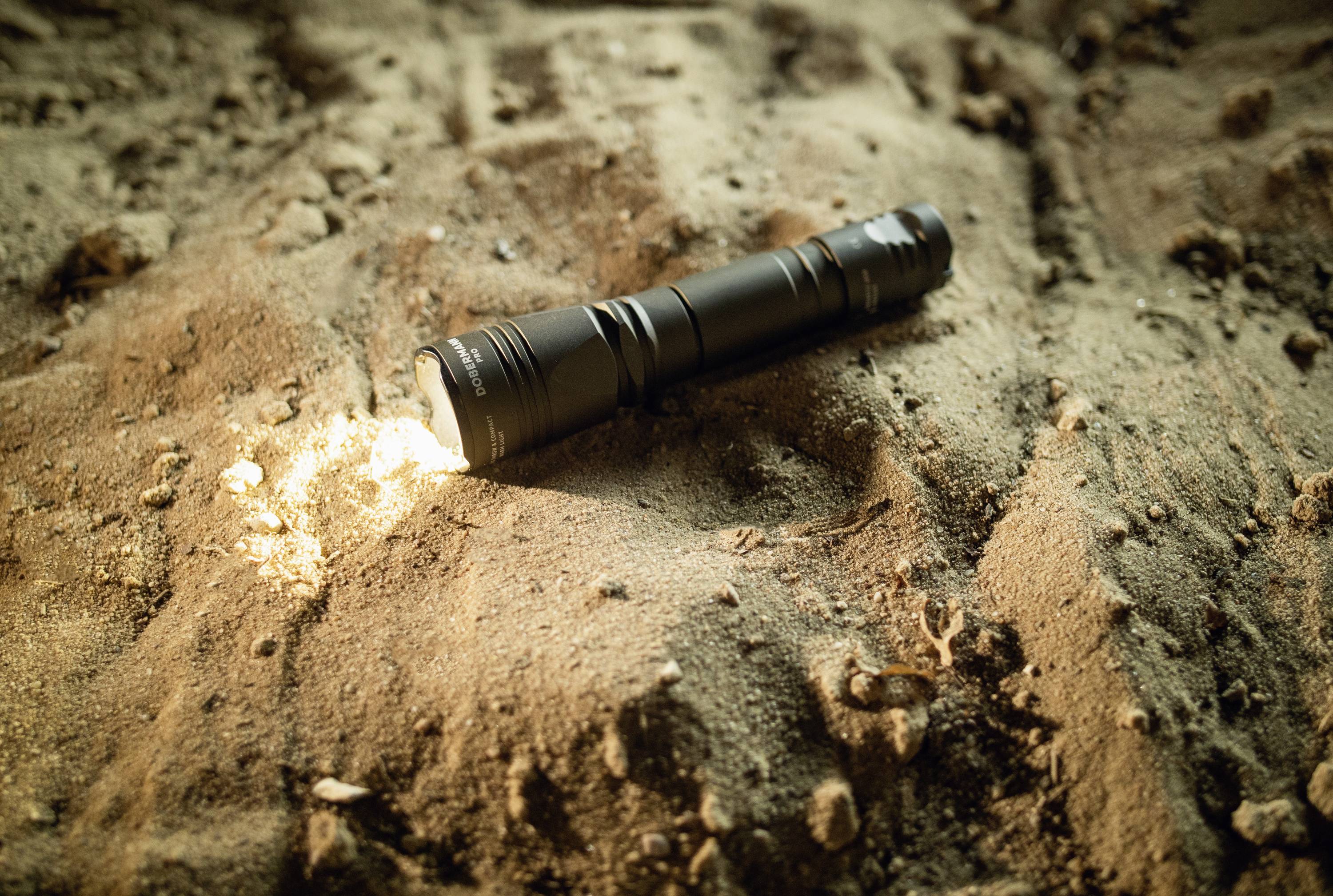 Taschenlampe liegt eingeschaltet auf sandigem Untergrund. Sie beleuchtet den Sand leicht und hinterlässt einen kleinen Lichtkreis.