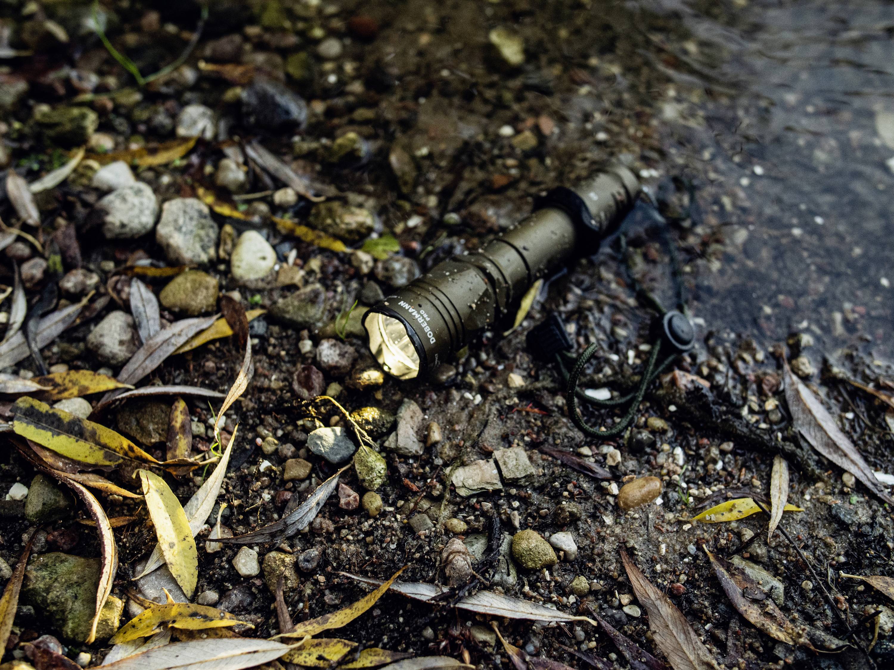 Taschenlampe liegt am Ufer eines Bachlaufs im Wald, umgeben von Steinen und nassem Laub. Die Lampe leuchtet, trotz Nässe.