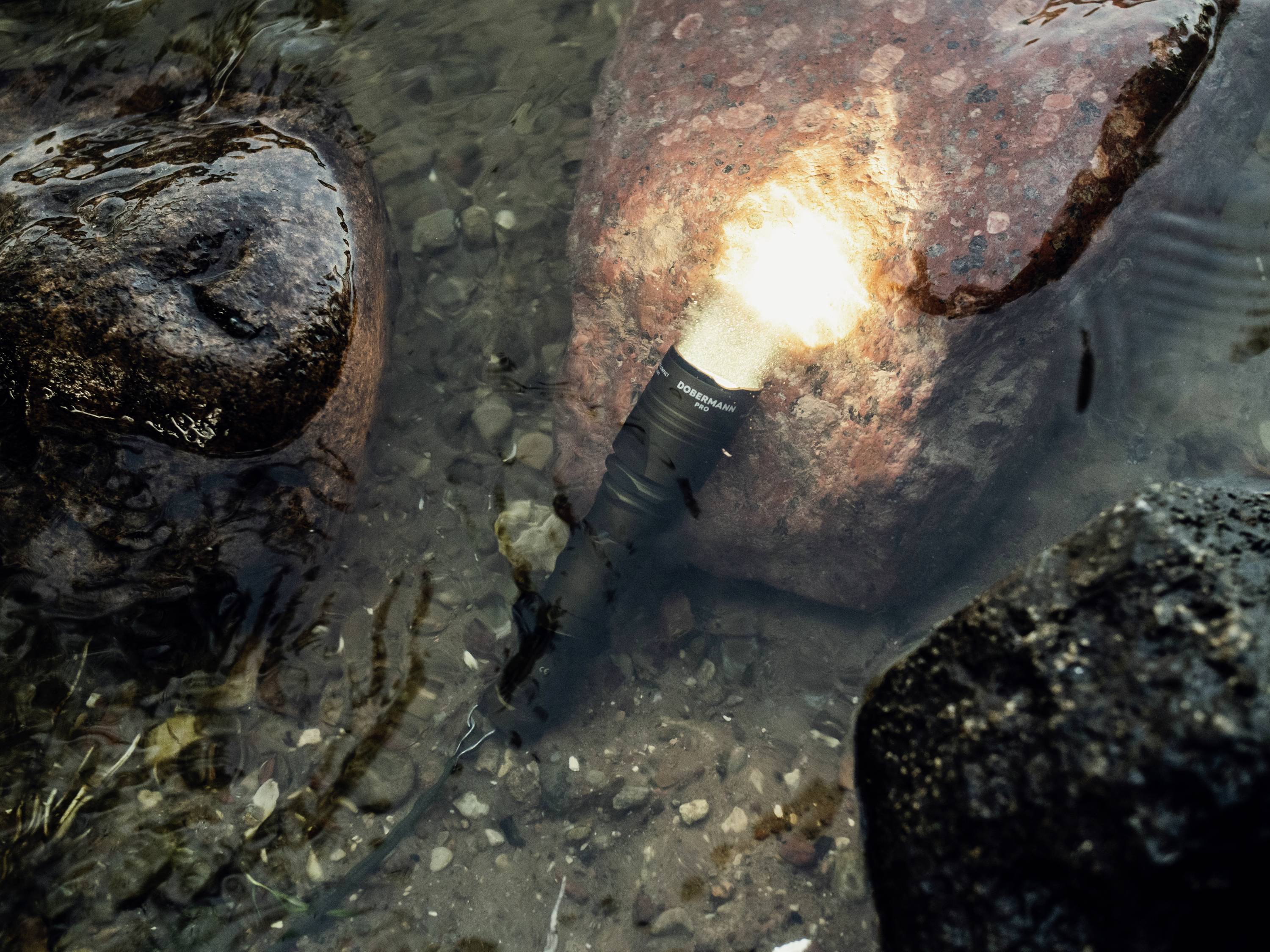 Unter Wasser strahlende Taschenlampe, neben mehreren Steinen in klarem Wasser liegend.
