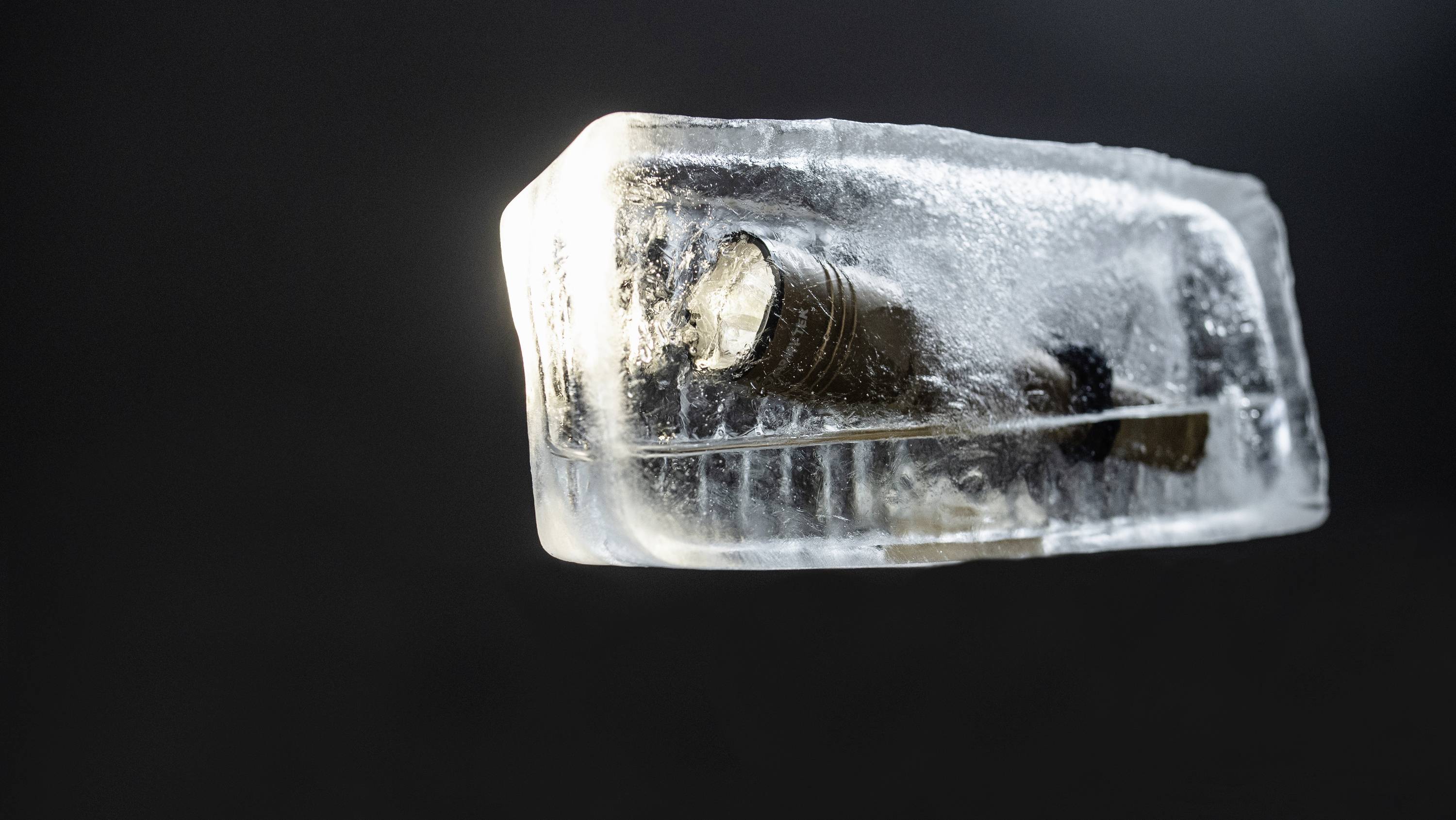 Gefrorene Taschenlampe eingebettet in Eisblock, vor dunklem Hintergrund. Lichtschein schimmert durch das Eis.
