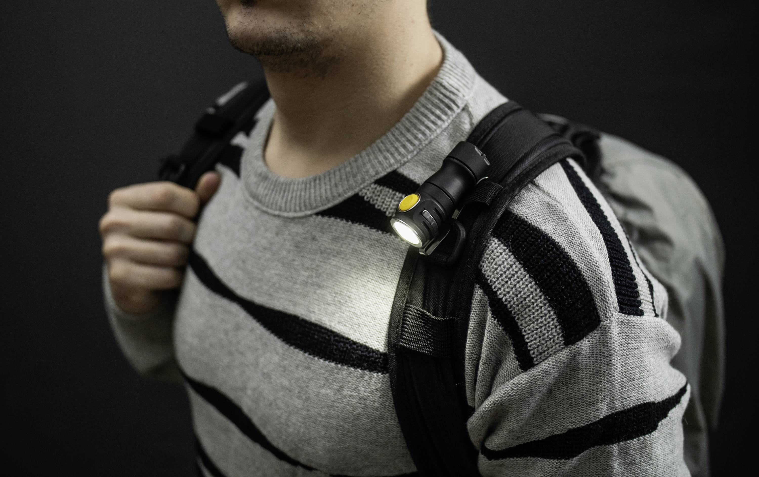 Eine Person trägt einen grauen Rucksack mit einem leuchtenden, an der Schulter befestigten kleinen Licht in einem dunklen Hintergrund.