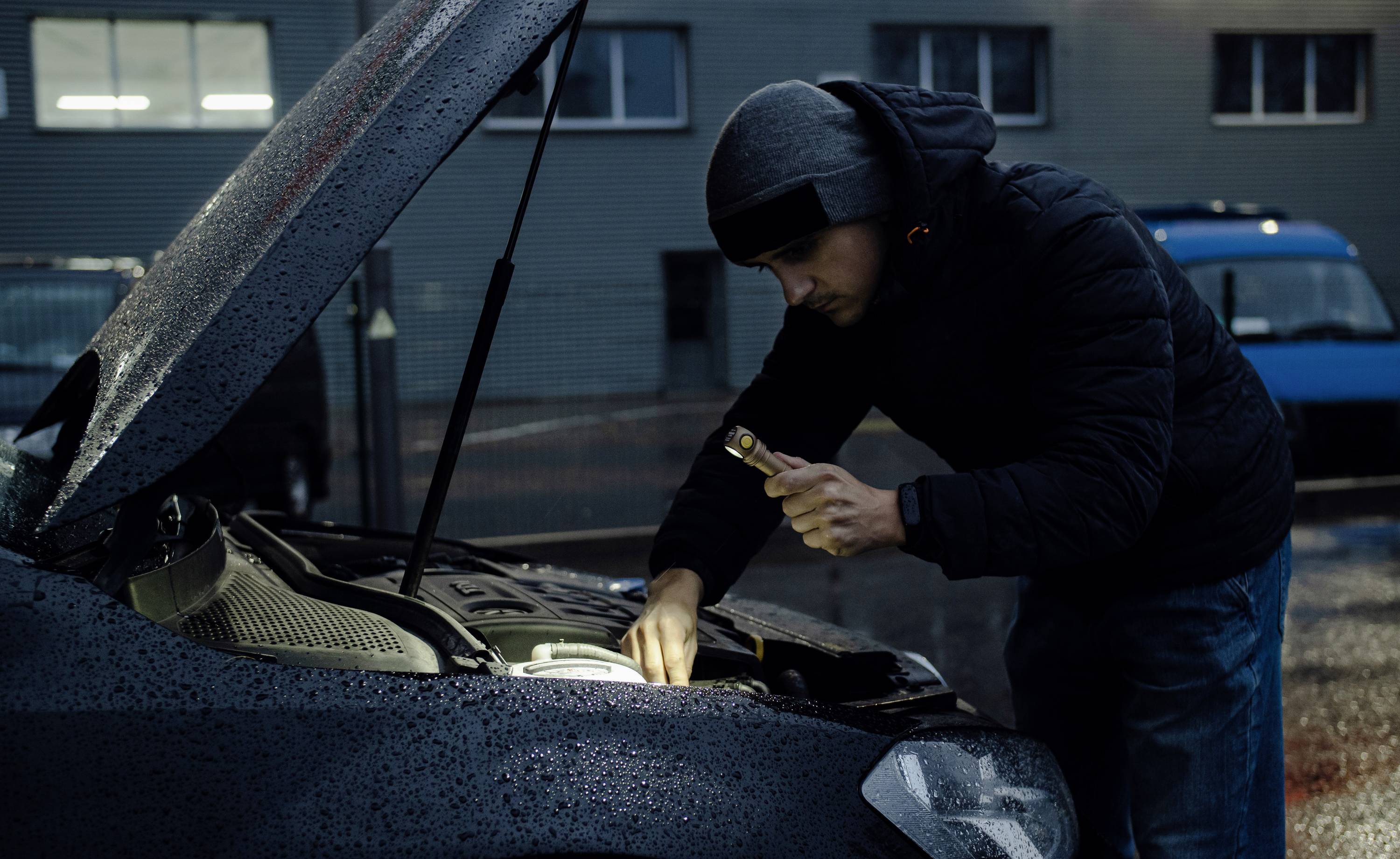 Eine Person im Kapuzenpullover beleuchtet mit einer Taschenlampe den Motorraum eines Autos, dessen Haube geöffnet ist. Es ist dunkel und regnerisch.