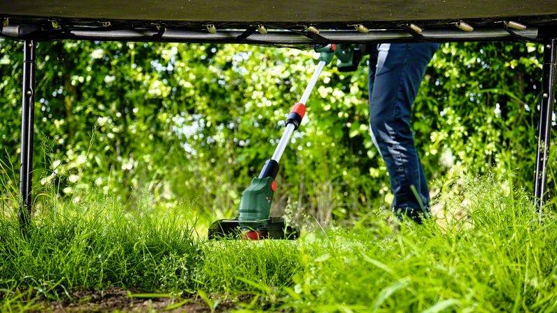 Eine Person mäht mit einem elektrischen Trimmer das hohe Gras unter einem Trampolin. Im Hintergrund sind grüne Büsche sichtbar.