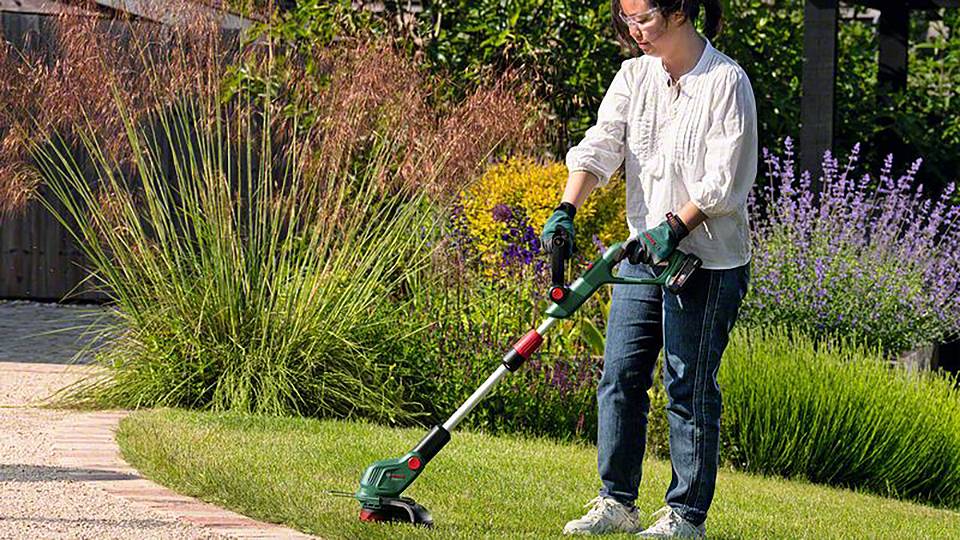 Person schneidet Rasen mit einem elektrischen Rasentrimmer im Garten. Umgeben von bunten Blumen und grünen Büschen. Wetter ist sonnig.