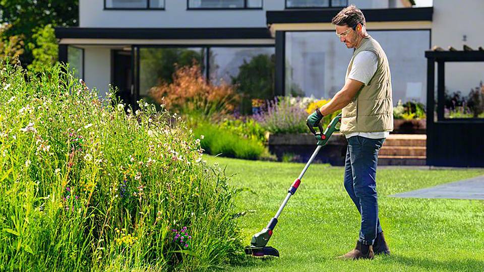 Ein Mann verwendet einen Rasentrimmer, um Gras an der Kante eines gepflegten Gartens zu schneiden. Im Hintergrund steht ein modernes Haus.