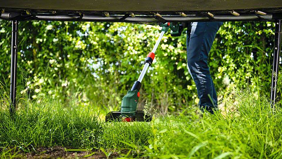 Mensch mäht Gras unter einem Trampolin mit einem elektrischen Rasentrimmer; grüne Büsche im Hintergrund.