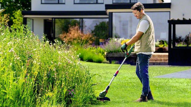 Ein Mann mäht hohes Gras mit einem Rasentrimmer vor einem modernen Haus. Er trägt Handschuhe und eine Weste. Im Hintergrund ist ein Garten.