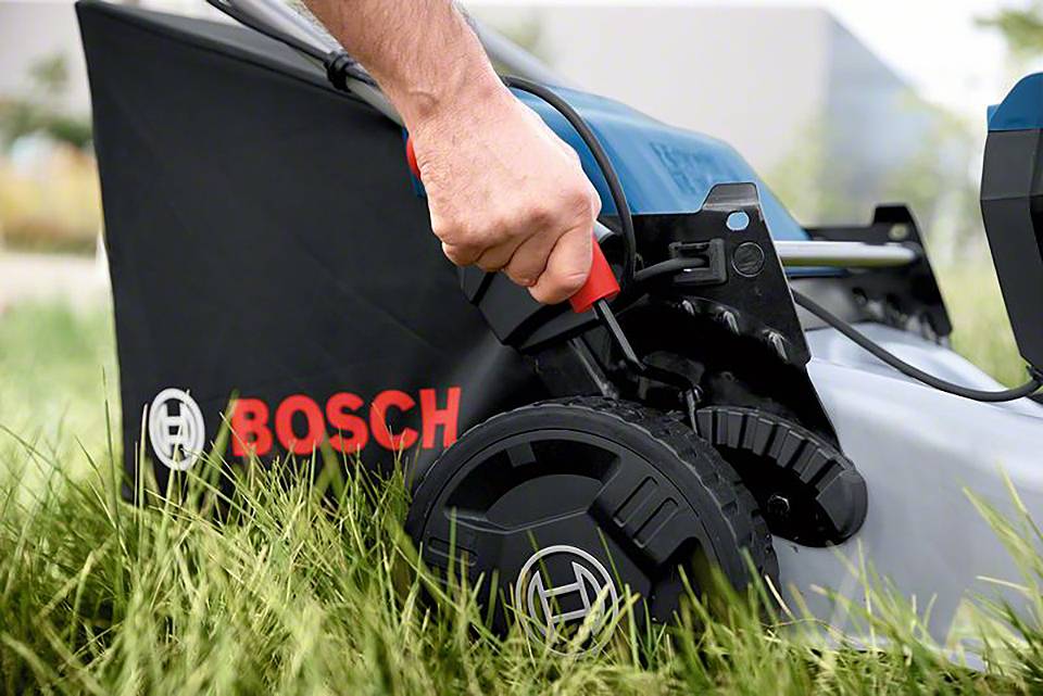 Eine Person passt die Schnitthöhe eines Bosch-Rasenmähers an einem Rad im hohen Gras an.