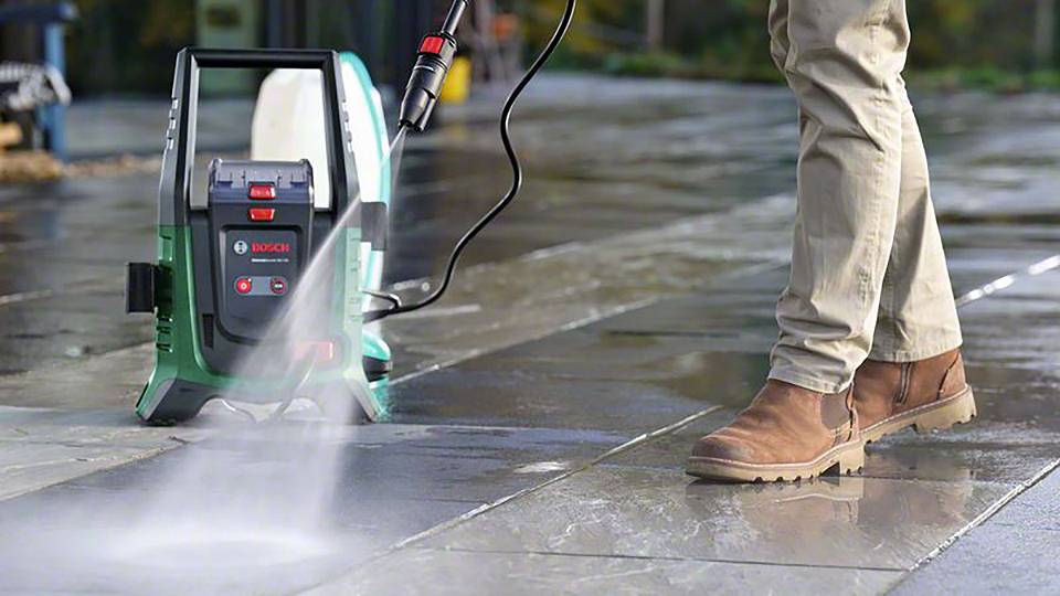Eine Person reinigt eine Terrasse mit einem Hochdruckreiniger. Die Nässe hebt den Schmutz vom Boden. Der Hochdruckreiniger ist grün.