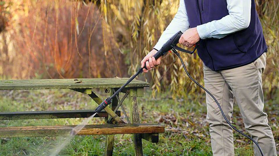 Eine Person reinigt mit einem Hochdruckreiniger eine hölzerne Bank im Freien. Im Hintergrund sind Bäume und Gras zu sehen.