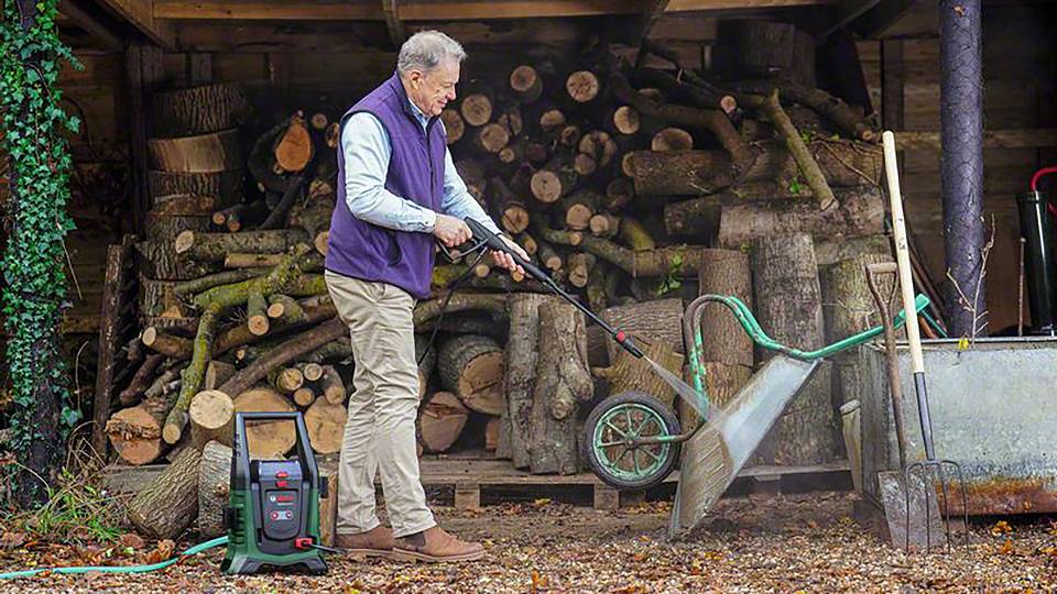 Ein Mann reinigt mit einem Hochdruckreiniger einen Behälter vor einem Stapel Holzscheite.