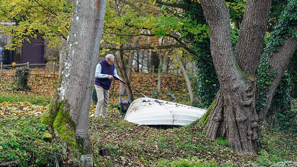 Älterer Mann im Park rechen Herbstlaub mit Harke; umgeben von großen Bäumen, Laub in herbstlichen Farben auf dem Boden verteilt.