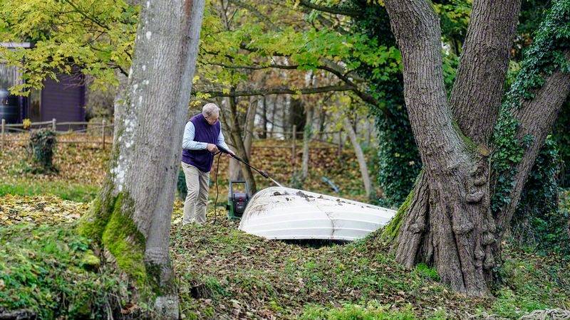 Eine Person harkt Laub rund um ein umgedrehtes Ruderboot im herbstlichen Garten, umgeben von bunten Bäumen und Grasflächen.