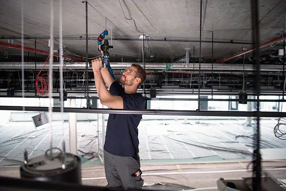 Eine Person installiert ein Gerät an der Decke eines Gebäudes. Der Raum wirkt unfertig, mit sichtbaren Leitungen und Rohren.