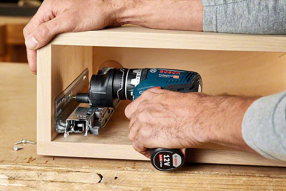 Person montiert eine Schublade mit einer elektrischen Bohrmaschine in einem Holzschrank, sichtbar sind Hände und Werkzeuge.