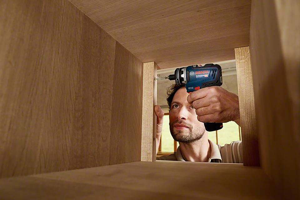 Ein Mann schraubt ein Holzregal zusammen. Er verwendet einen blauen Akkuschrauber und arbeitet konzentriert.