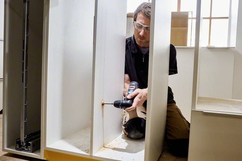 Ein Mann benutzt eine Bohrmaschine, um ein Möbelstück aus Holz zusammenzubauen. Er trägt Schutzbrille und kniet auf dem Boden.