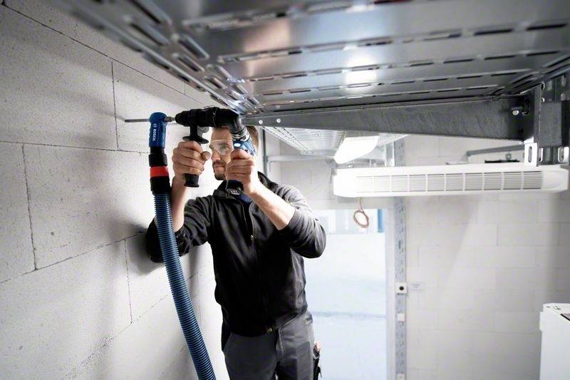 Eine Person installiert ein blaues Lüftungsrohr in einem technisch aussehenden Raum. Sie verwendet Werkzeug, um das Rohr zu befestigen.