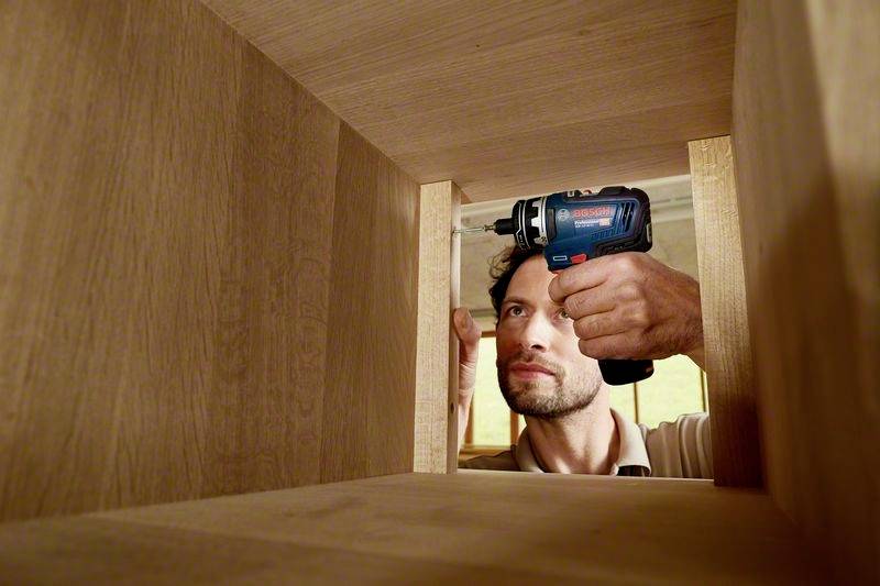 Ein Mann arbeitet mit einem elektrischen Schraubenzieher in einem Holzmöbelstück. Er befestigt Teile im Inneren des Möbels.