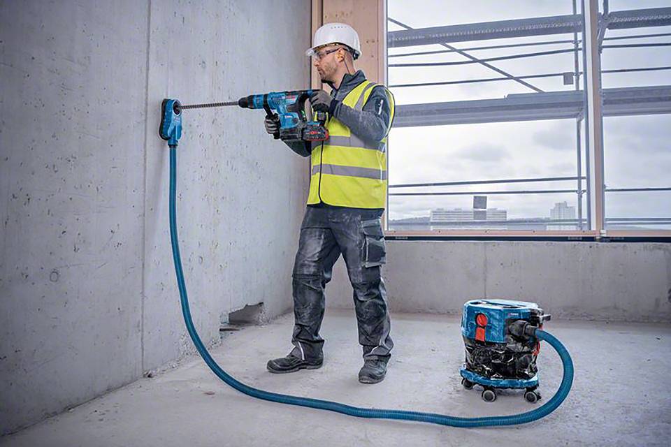 Ein Arbeiter mit Schutzhelm und Sicherheitsweste bohrt ein Loch in eine Betonwand auf einer Baustelle. Neben ihm steht ein Staubsauger.
