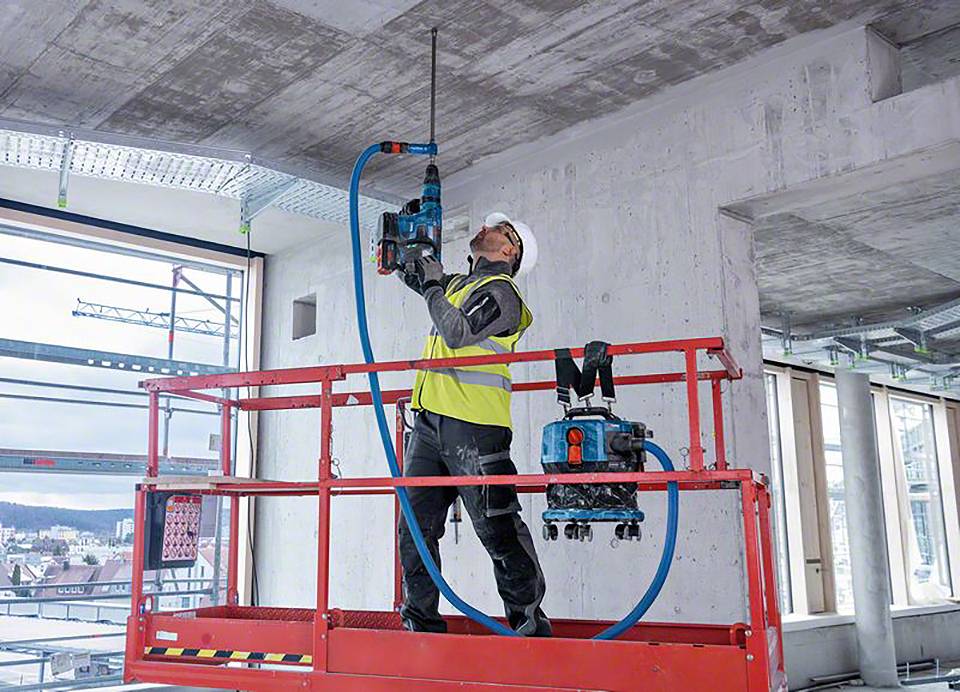 Arbeiter bohrt mit einer Maschine ein Loch in die Decke eines Gebäudes, steht auf einer Hebebühne, trägt Schutzkleidung und Helm.