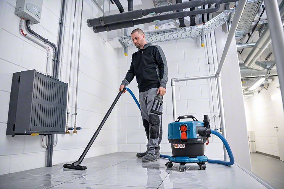 Ein Mann reinigt den Boden in einem Technikraum mit einem Industriesauger. Rohre und ein Verteilerkasten sind an der Wand sichtbar.